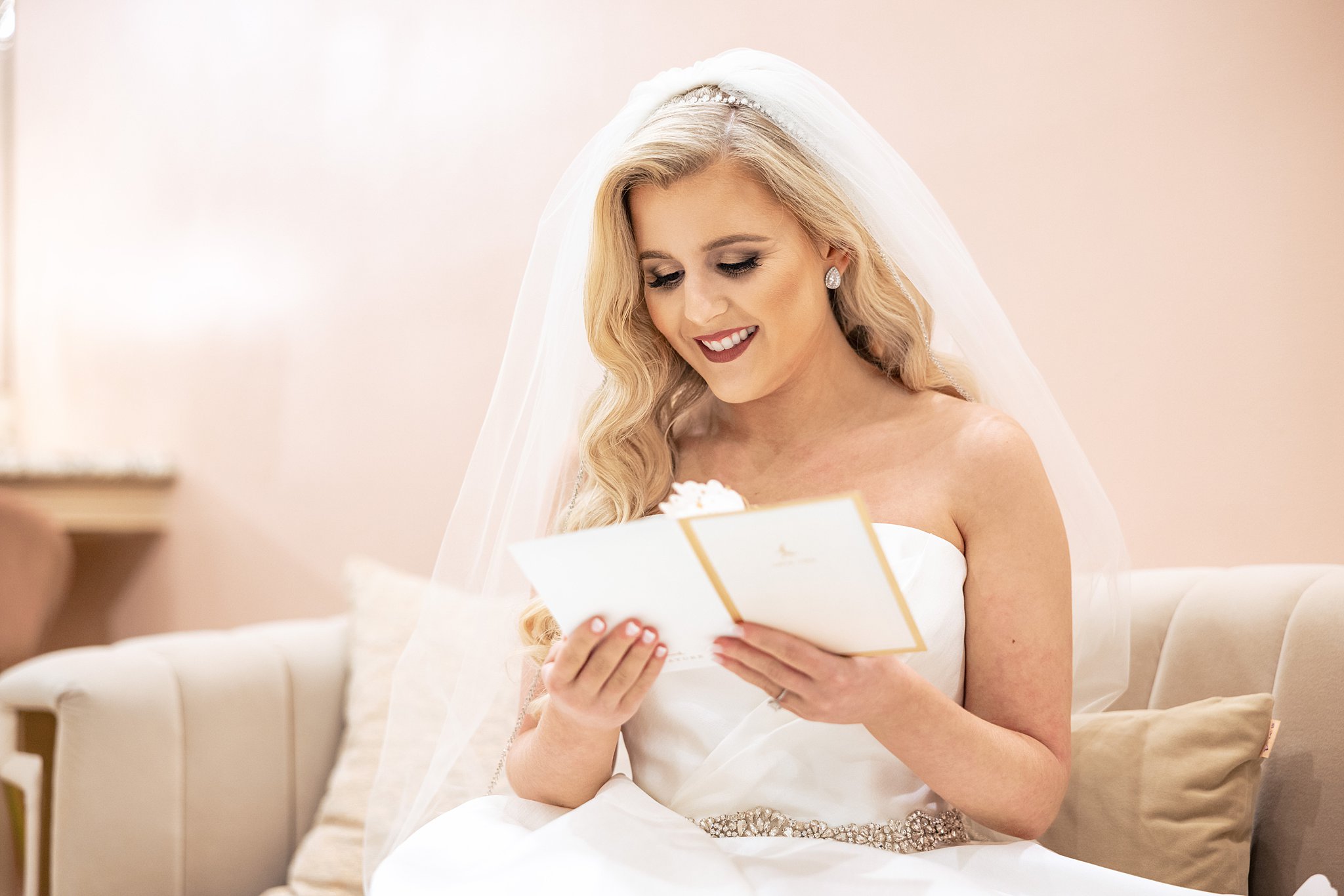 A bride sits on a couch in her dress and veil reading a card with a smile