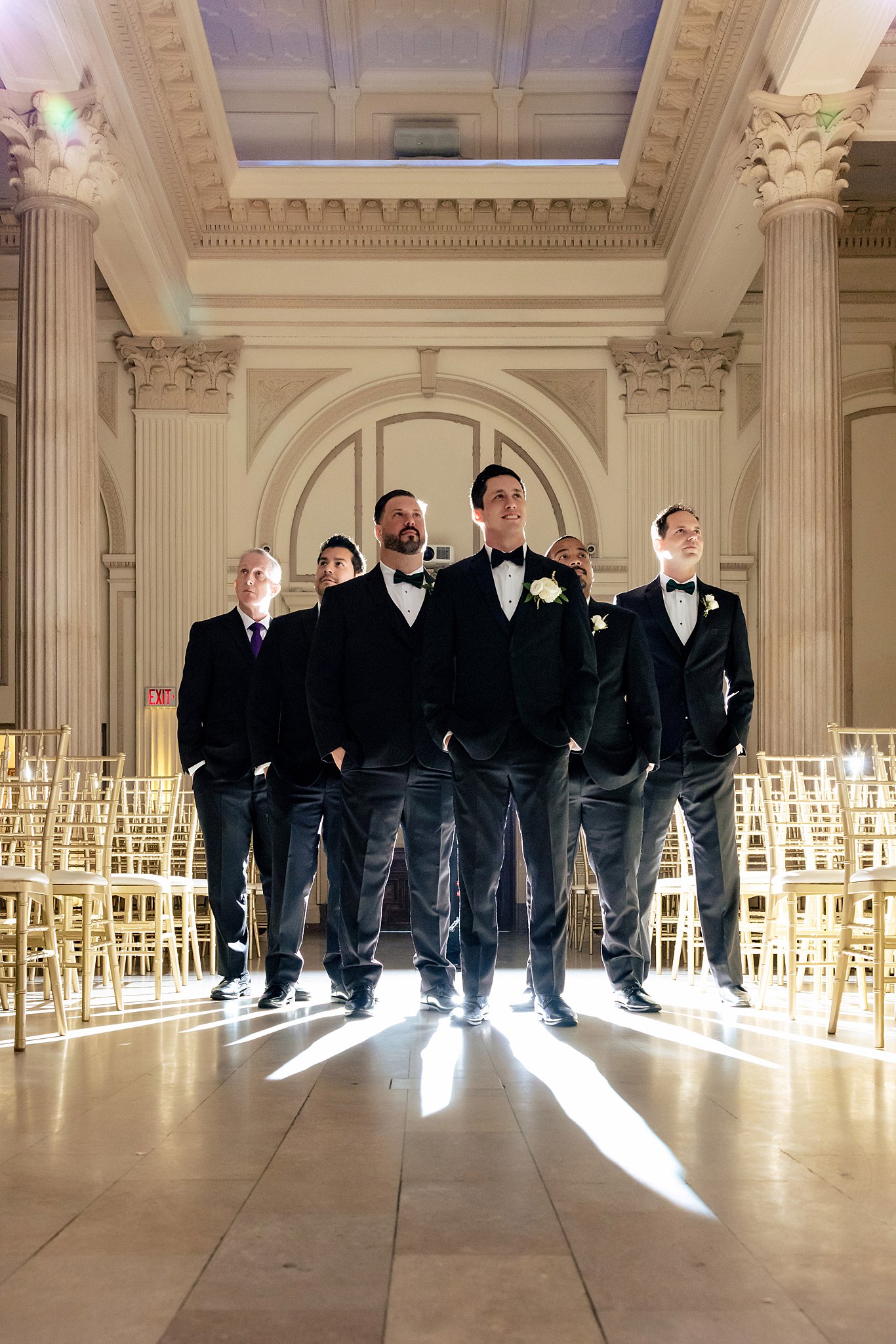A groom and his 5 groomsmen stand in an ornate wedding ceremony hall with hands in pockets