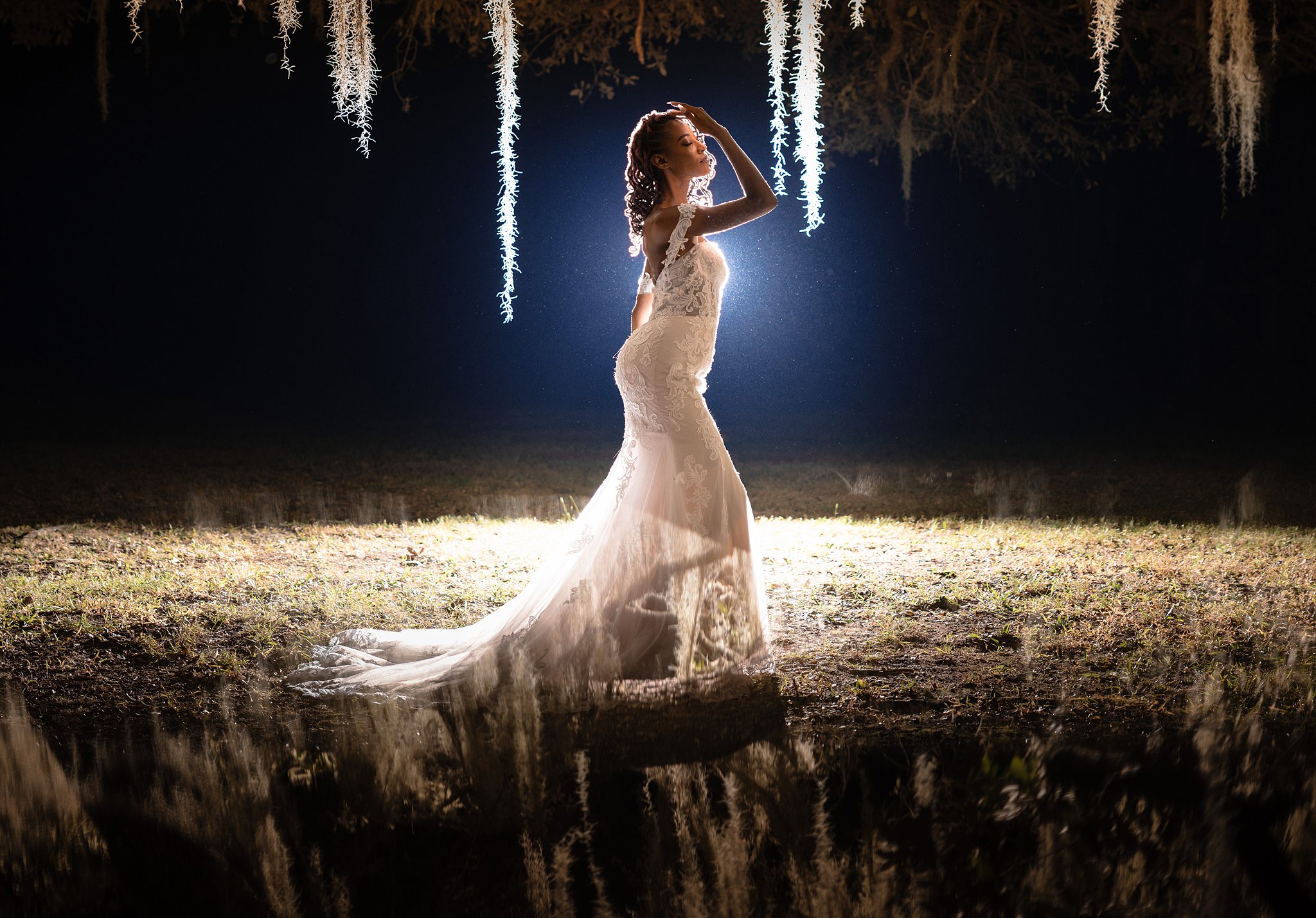 A bride stands in a pool of light in an embroidered lace dress with a hand reaching to her hair under an oak tree at one of the st augustine wedding venues