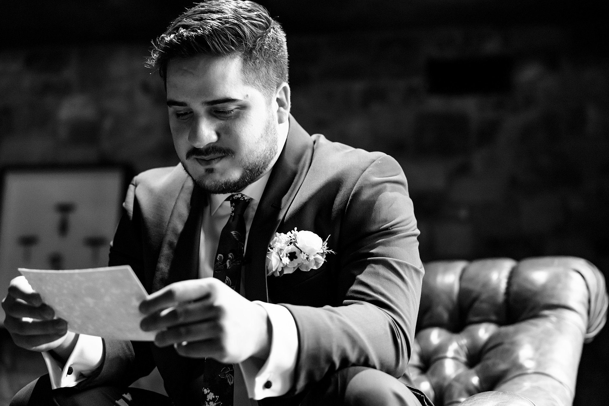 A groom sits on a leather chair reading a note from his bride before his ceremony