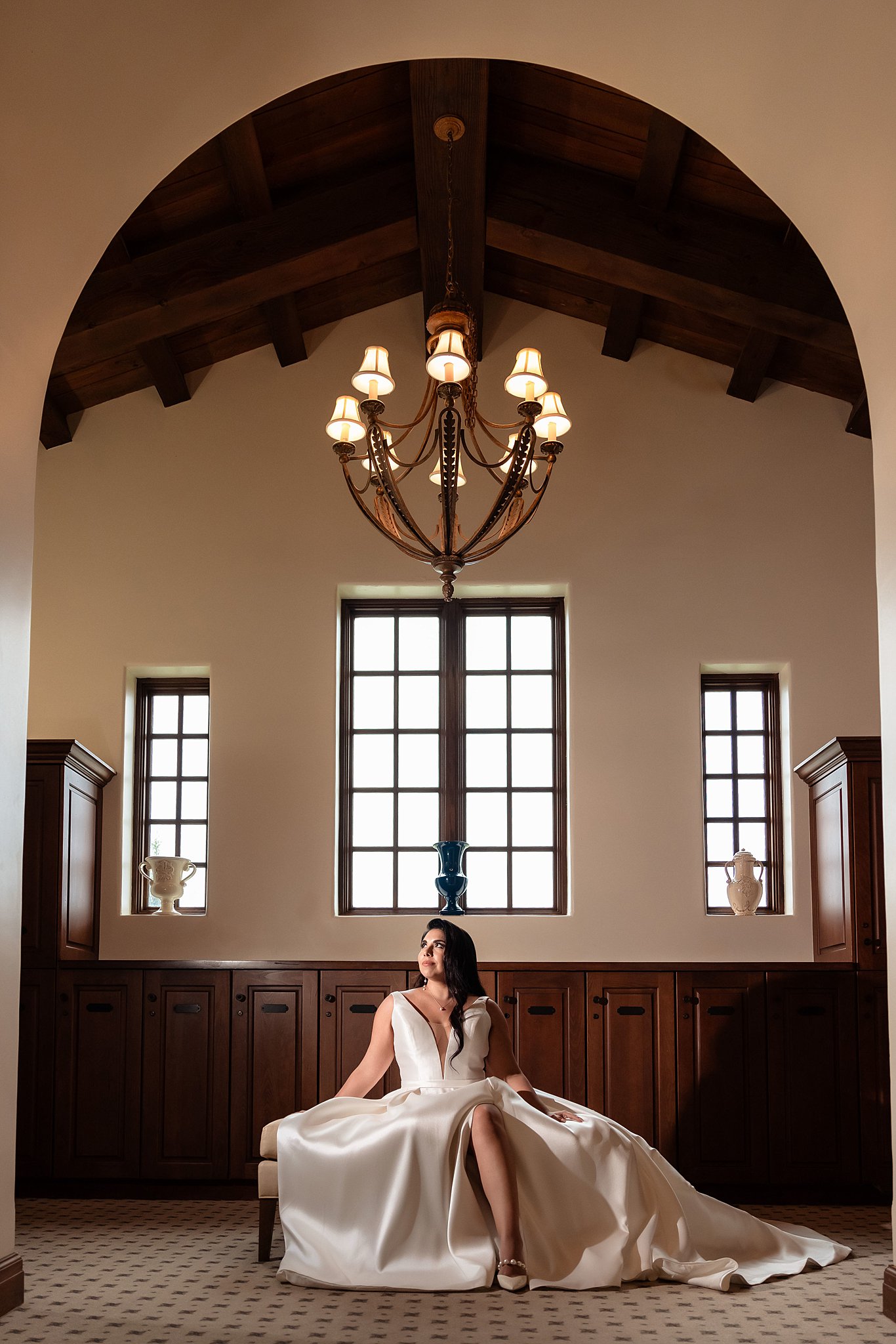 A bride sits on a bench in her ornate getting ready room in a silk dress