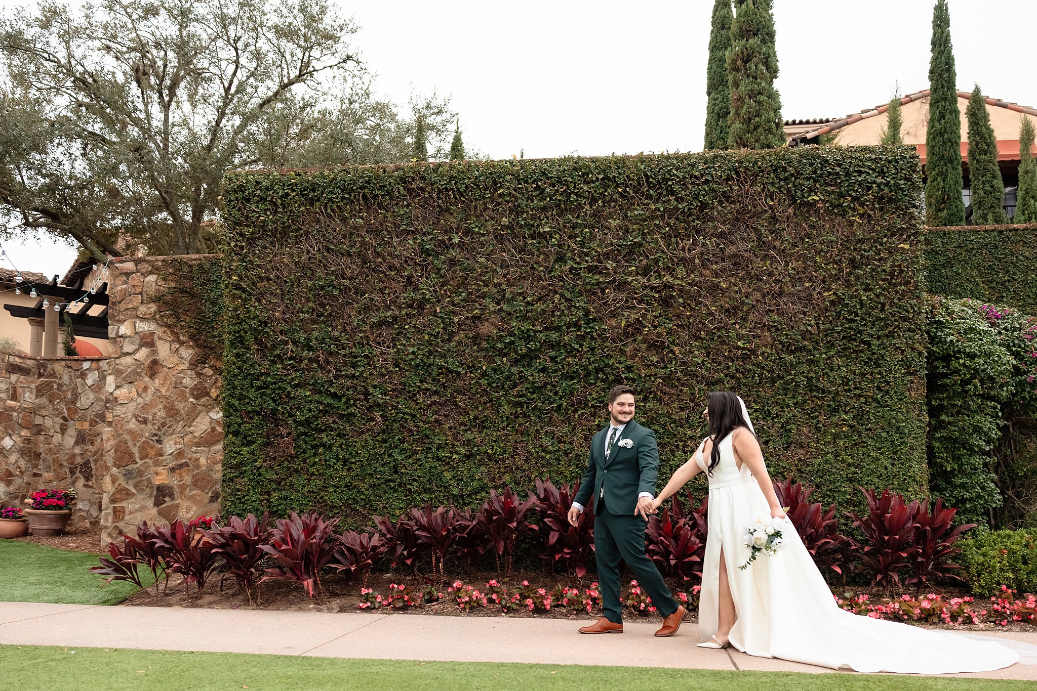 Newlyweds walk down a sidewalk through a garden and an ivy wall holding hands at their wedding