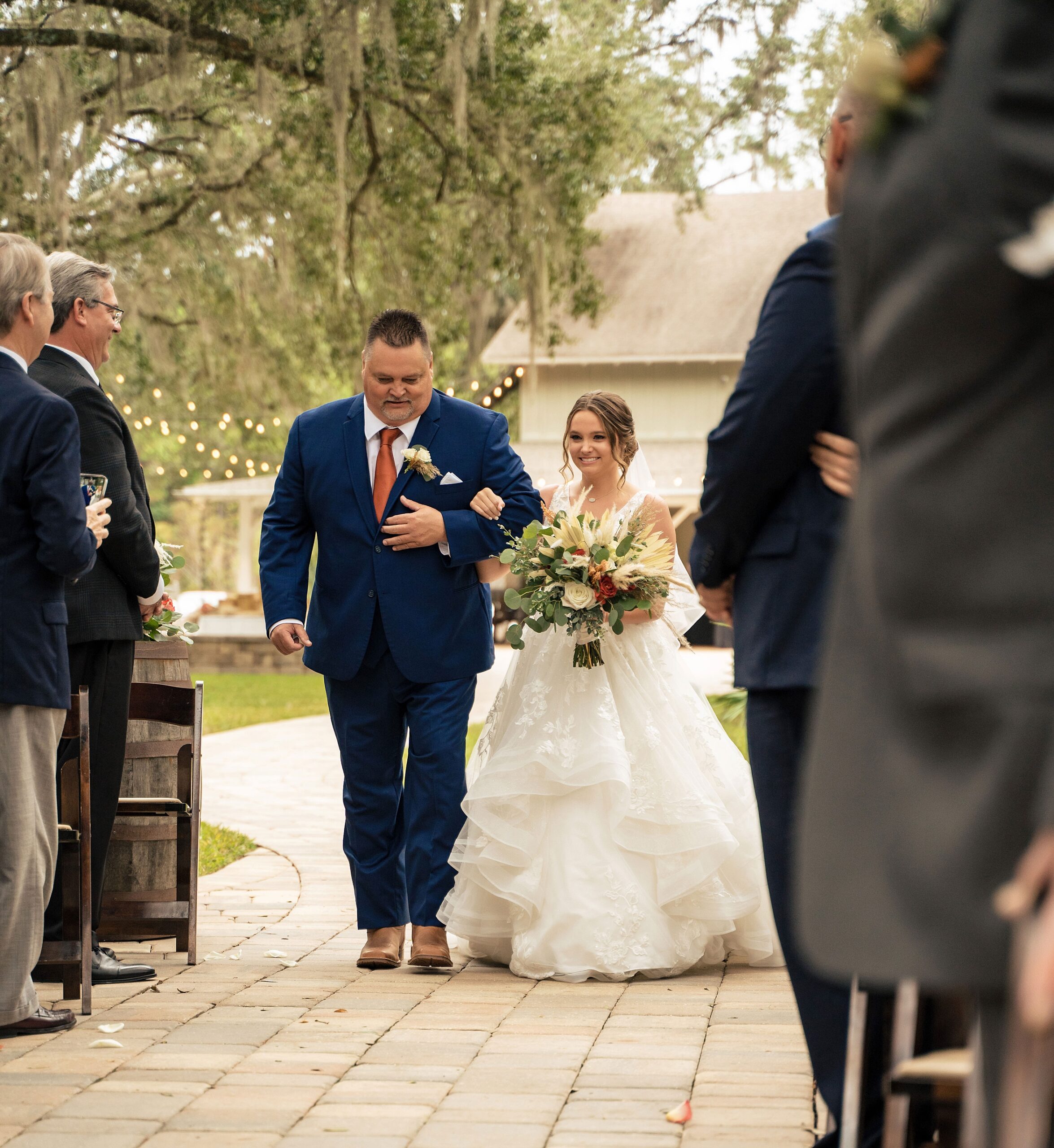 A father in a blue suit walks his daughter down the aisle of her outdoor patio wedding