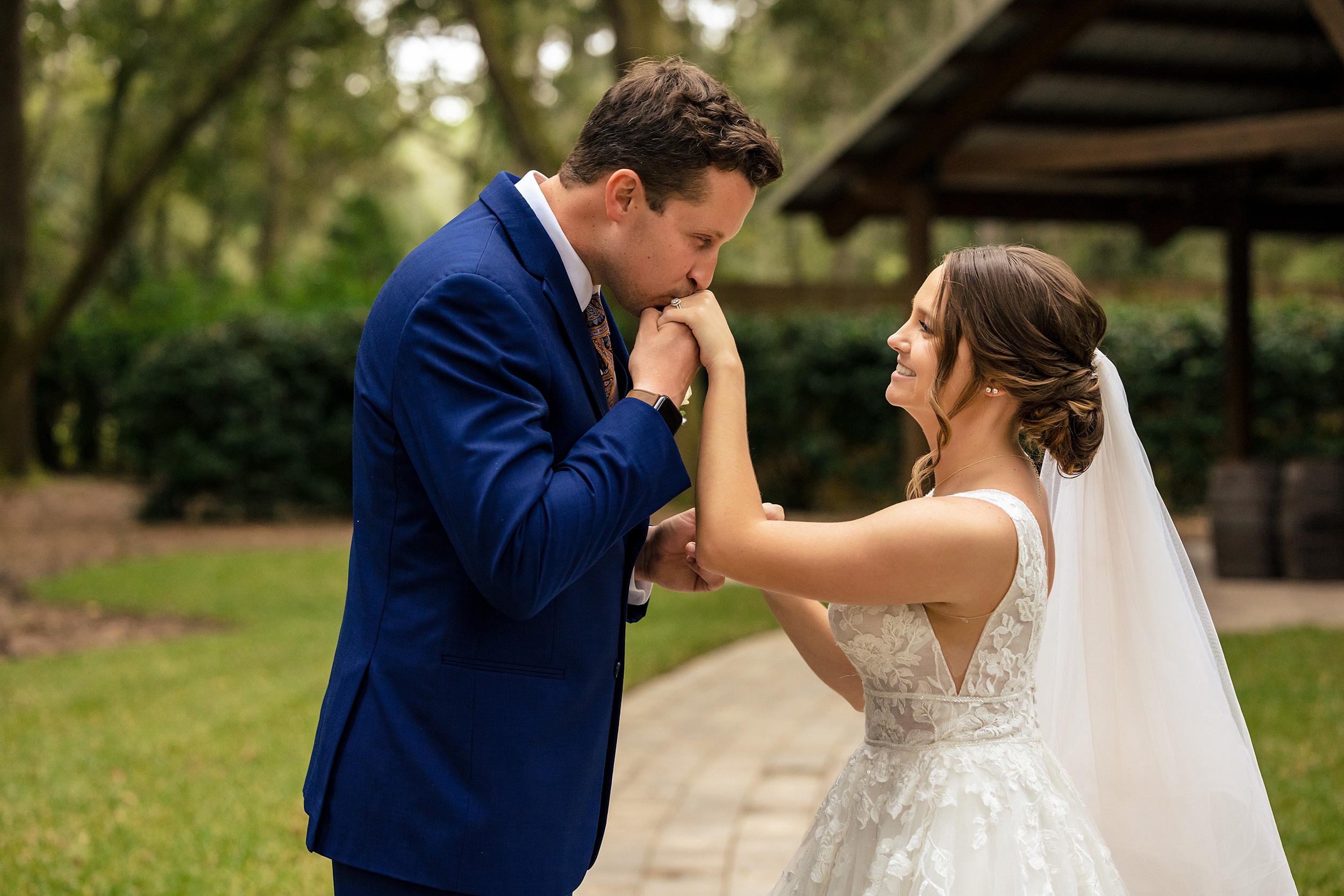 A groom in a blue suit kisses the hand of his bride in a lace dress during their first look