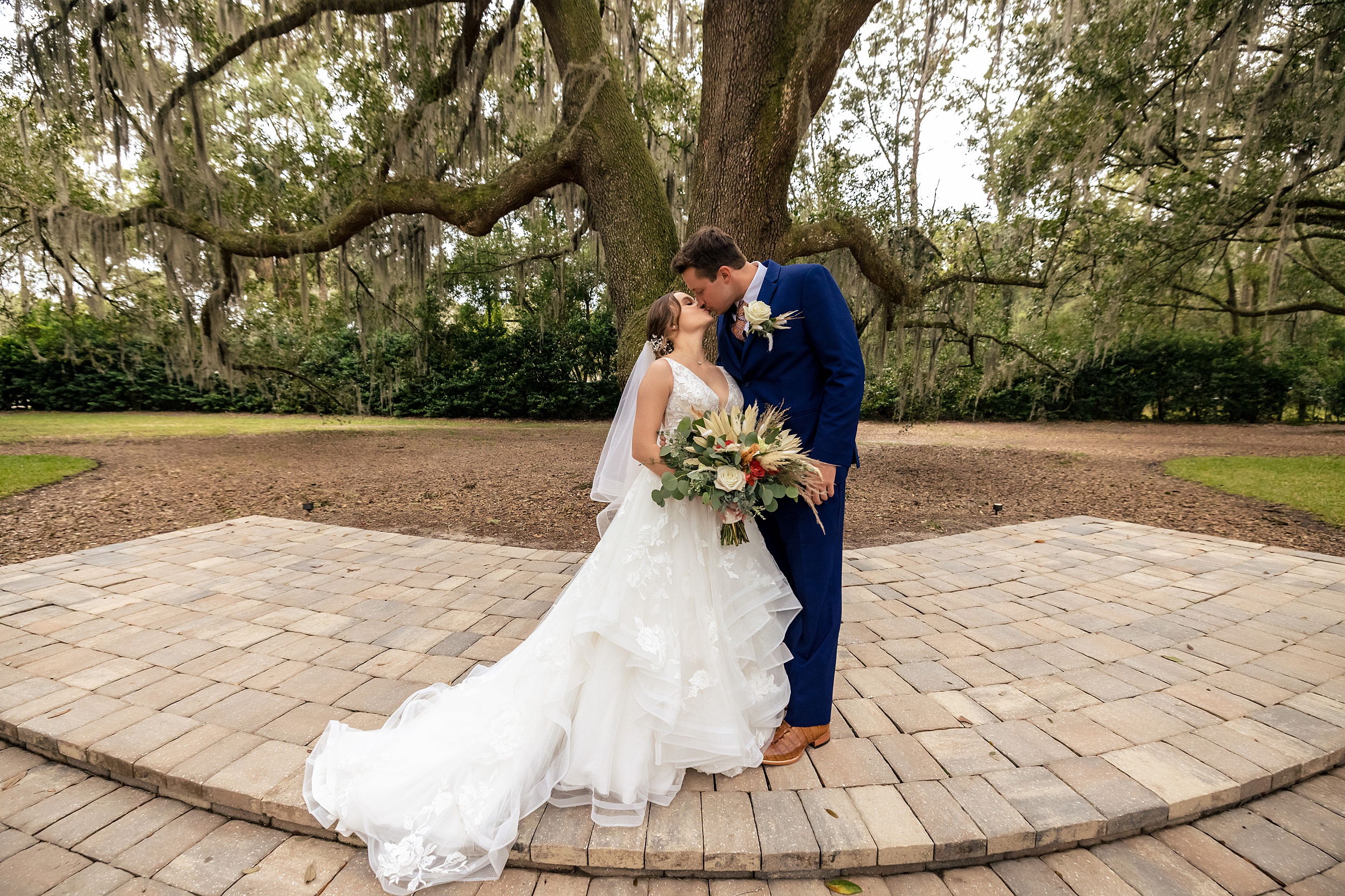 Newlyweds kiss while standing under a large oak tree on raised patio