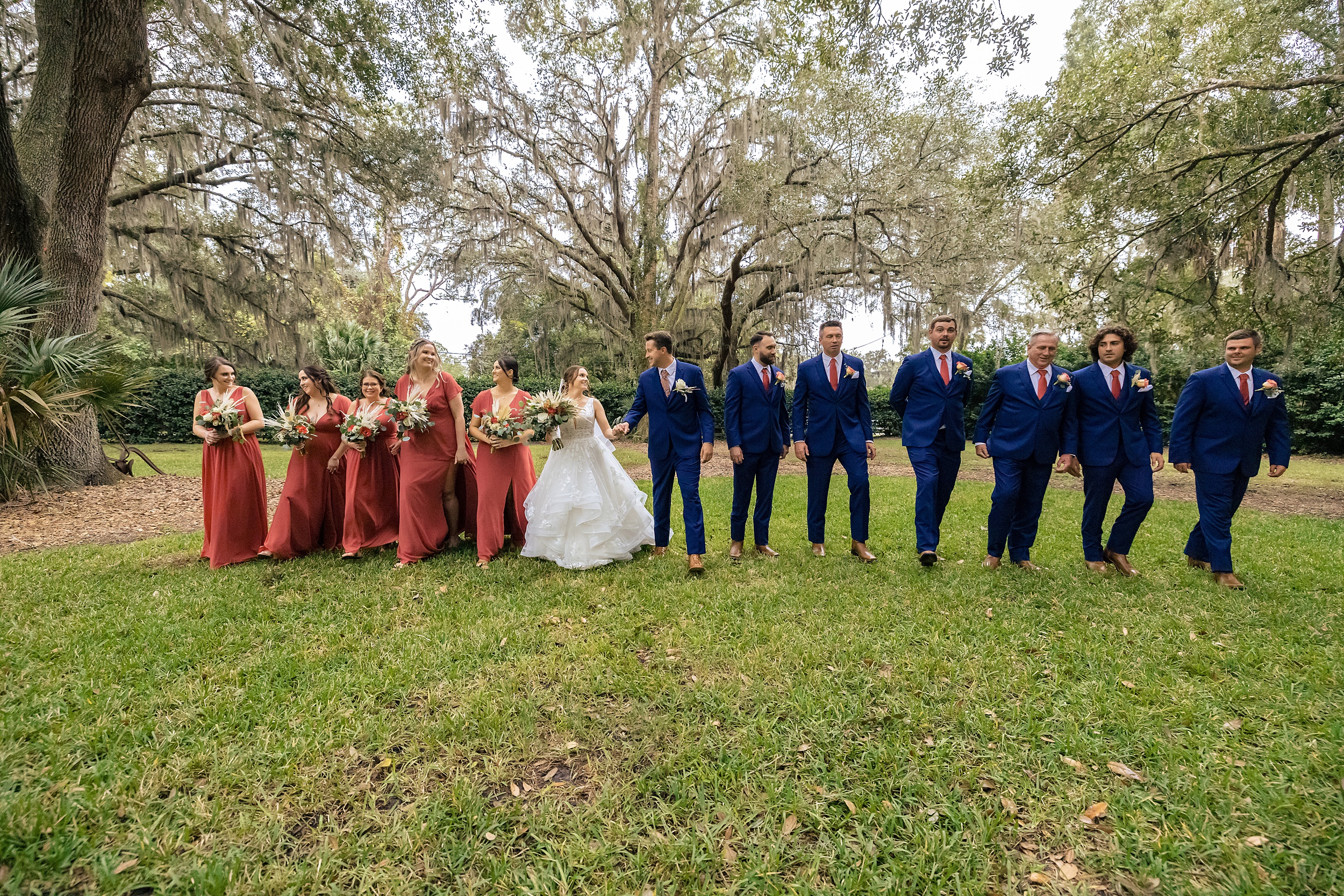 Newlyweds hold hands while walking through a grassy lawn under trees with their large wedding party at a bowing oaks wedding