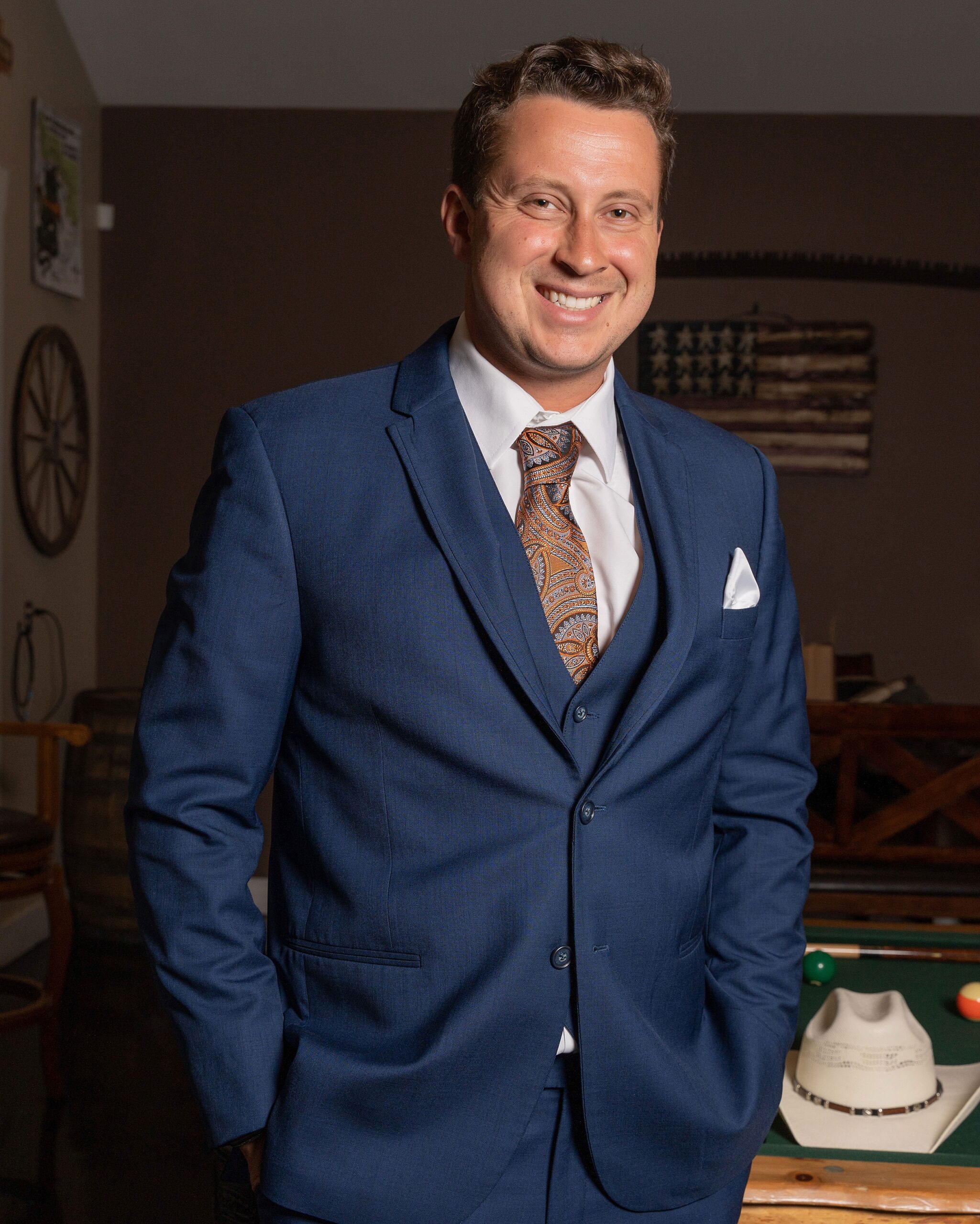 A groom stands by a pool table in a blue suit with hands in pockets