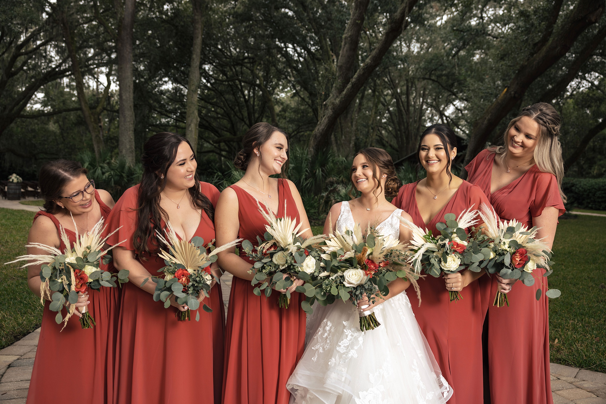 A bride laughs with her bridal party in red dresses holding their bouquets under large oak trees at her bowing oaks wedding