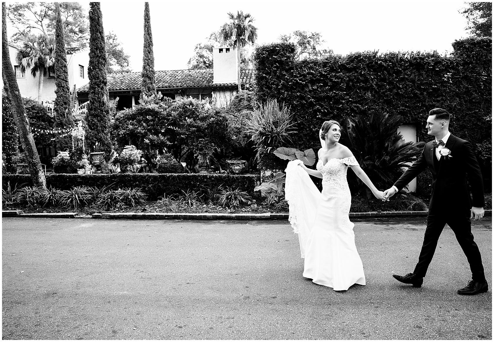 A bride leads her groom by the hand down a sidewalk through a garden