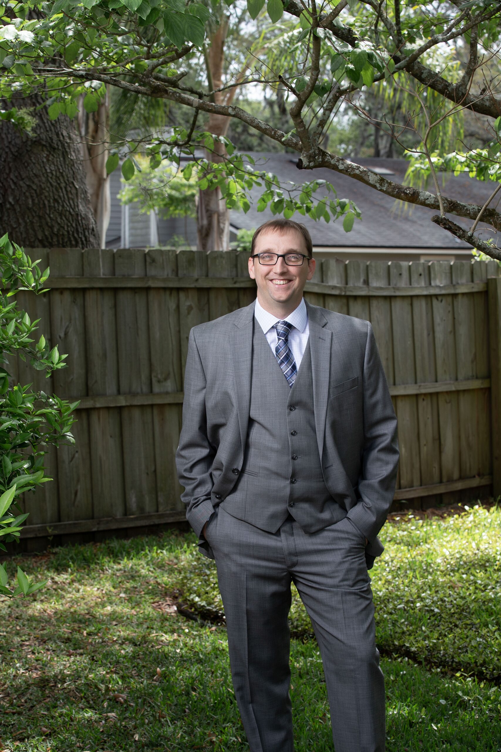 A groom in a grey suit stands in a yard with hands in pockets