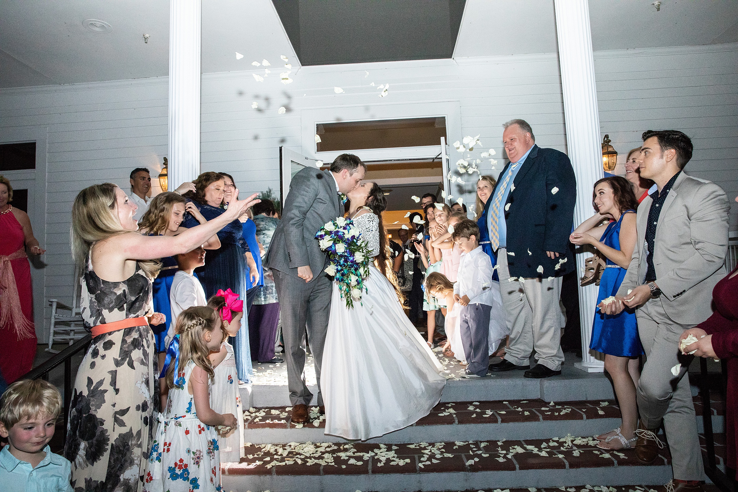 Newlyweds kiss at the steps of their wedding reception venue while leaving surrounded by guests throwing rose petals