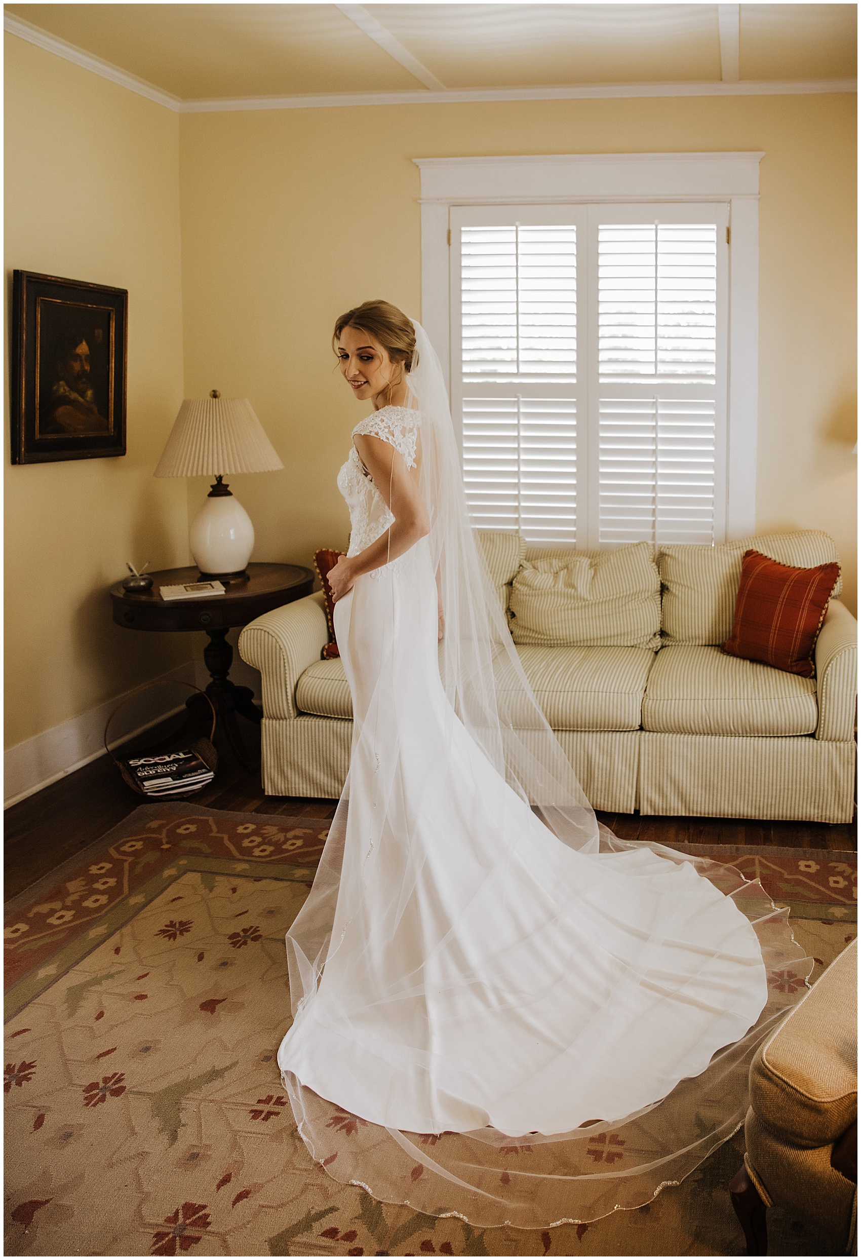 A bride stands in the getting ready room smiling over her shoulder to her train and veil stretched out