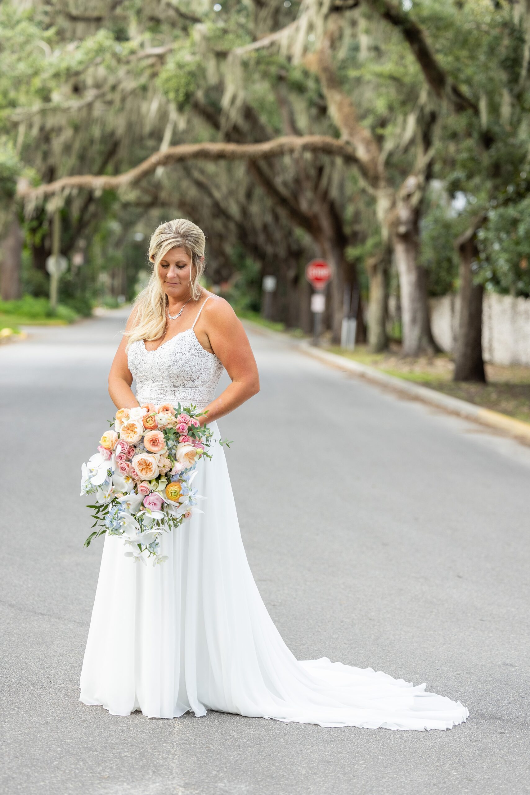 A bride smiles down at her colorful bouquet while standing in a street lined with oak trees at her fountain of youth wedding