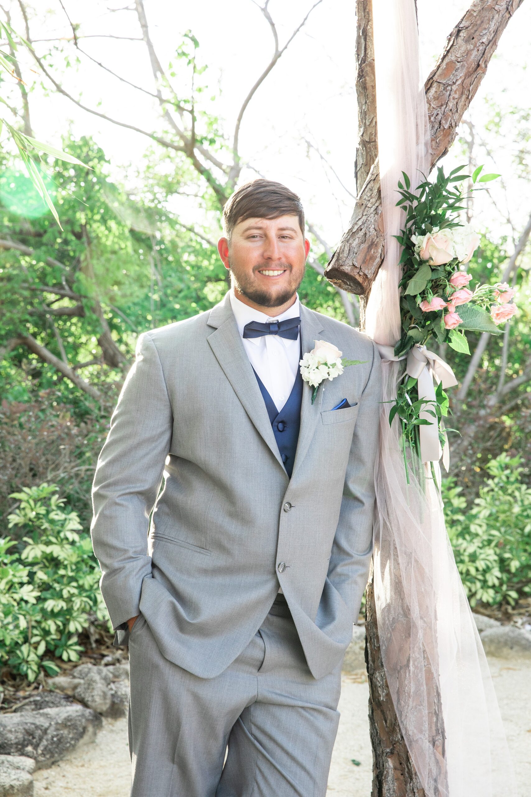 A groom in a grey suit leans against a pine tree arbor with pink flowers