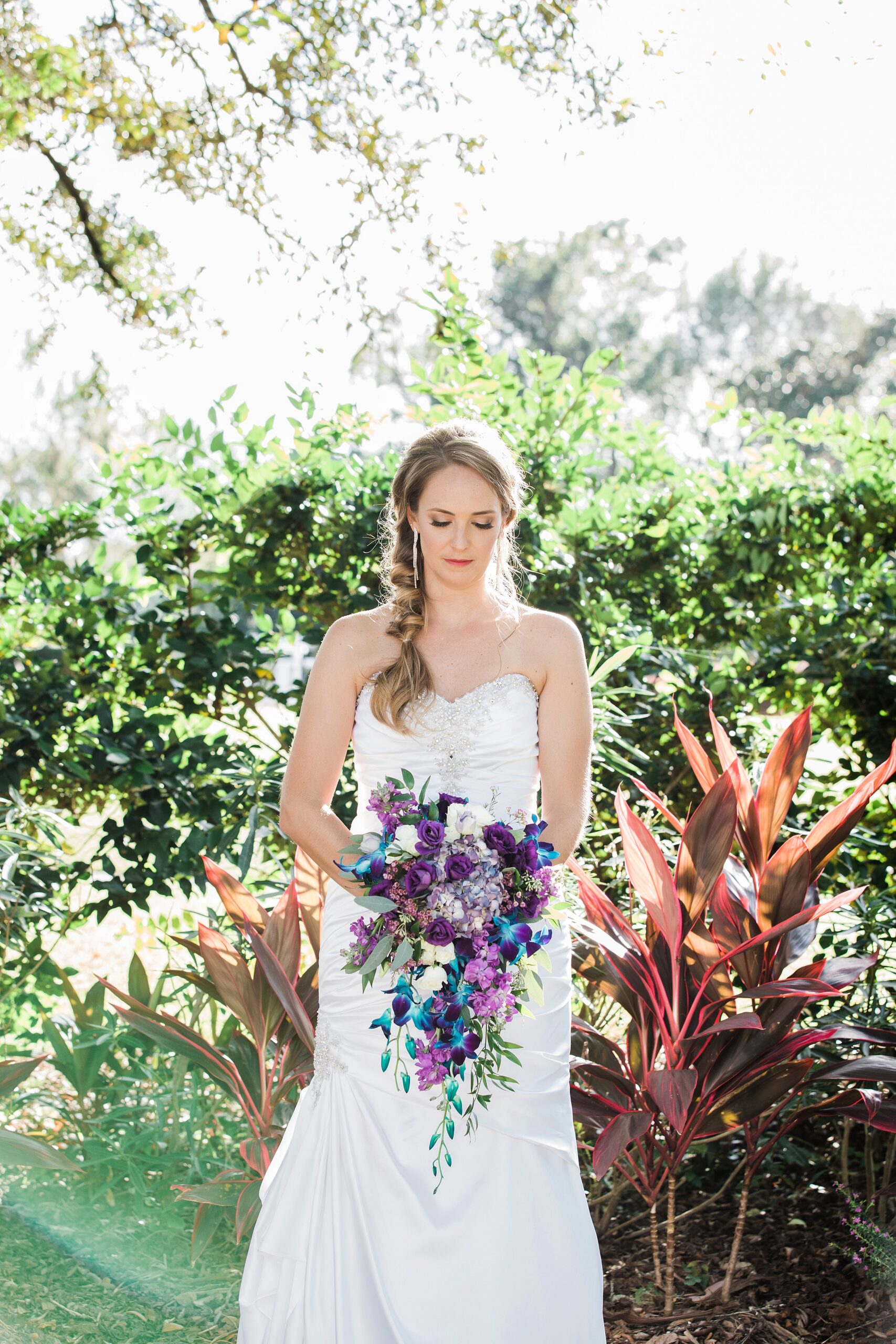 A bride looks down at her colorful blue and purple bouquet in a garden at her magnolia point wedding