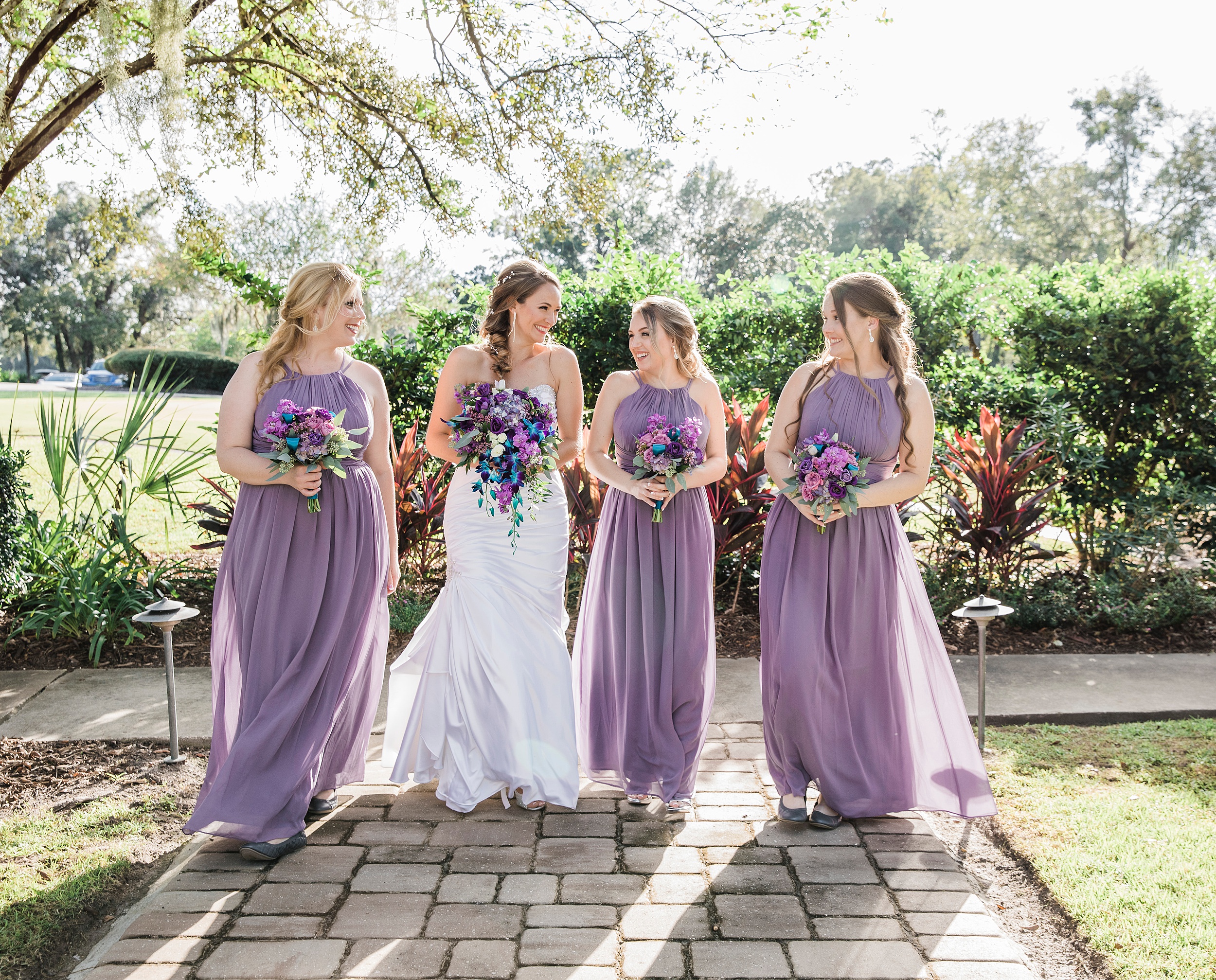 A bride laughs with her bridesmaids in purple dresses while walking through a garden path