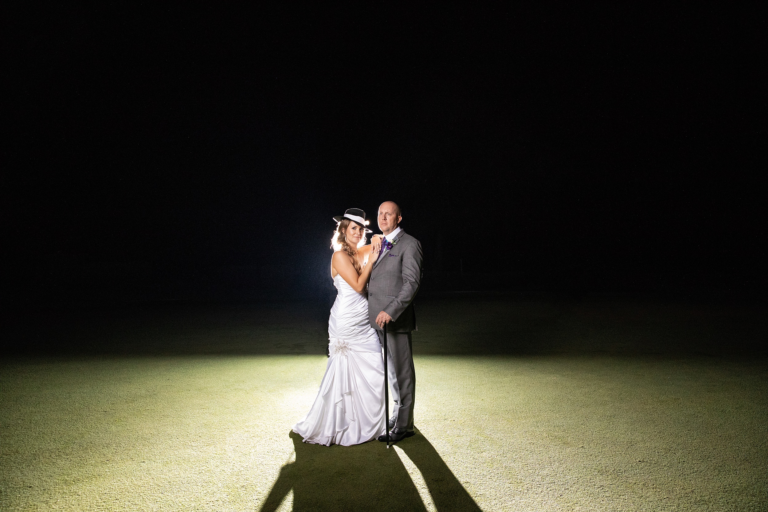 Newlyweds stand on a manicured lawn at night holding a cane and the bride wearing a hat