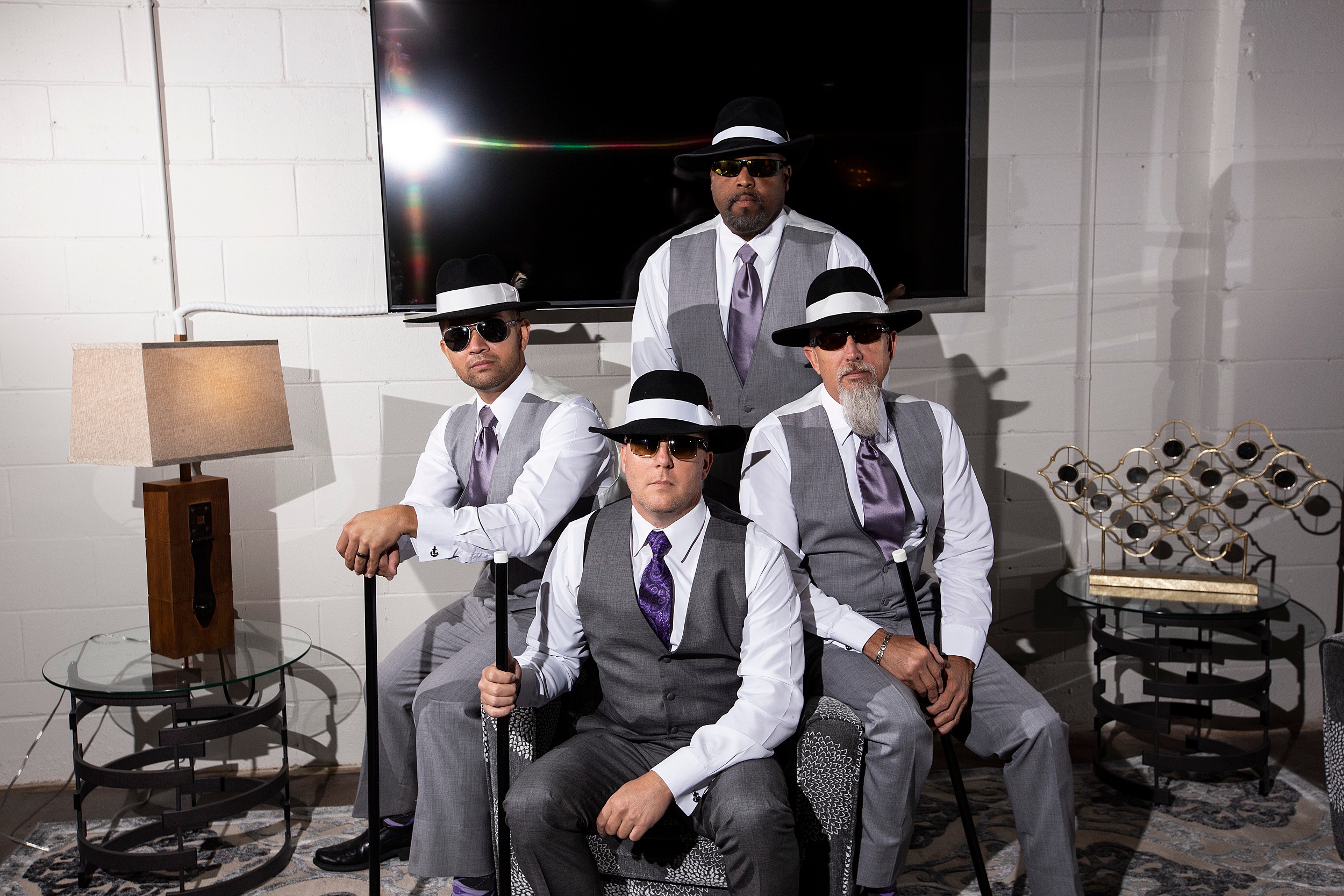 A groom sits in a modern style room with his groomsmen in large hats and holding canes with purple ties
