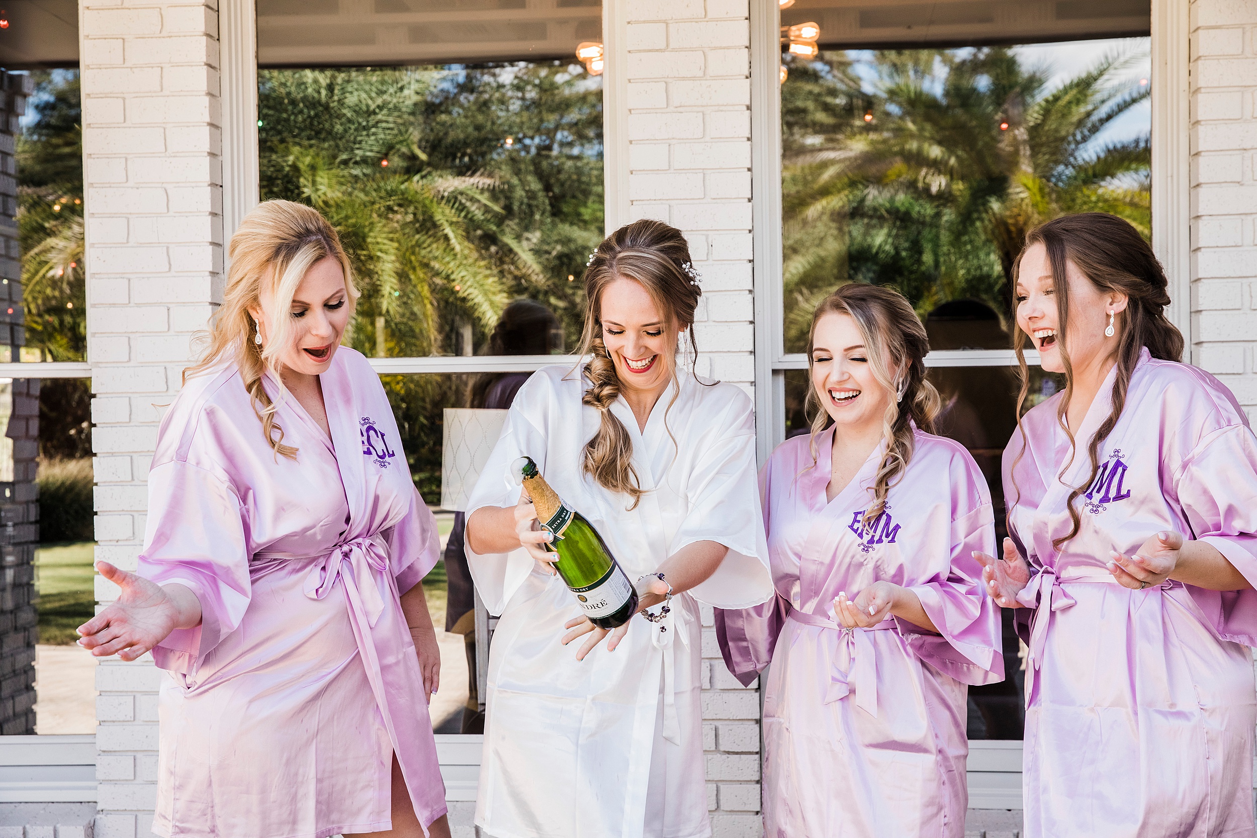 A bride in white pajamas pops champagne with her bridesmaids in pink pajamas