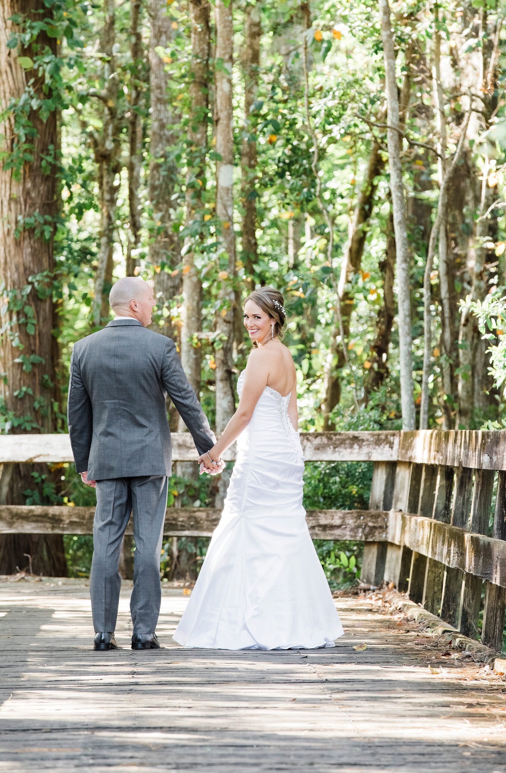 A bride stands on a boardwalk looking over her shoulder while holding hands with her husband