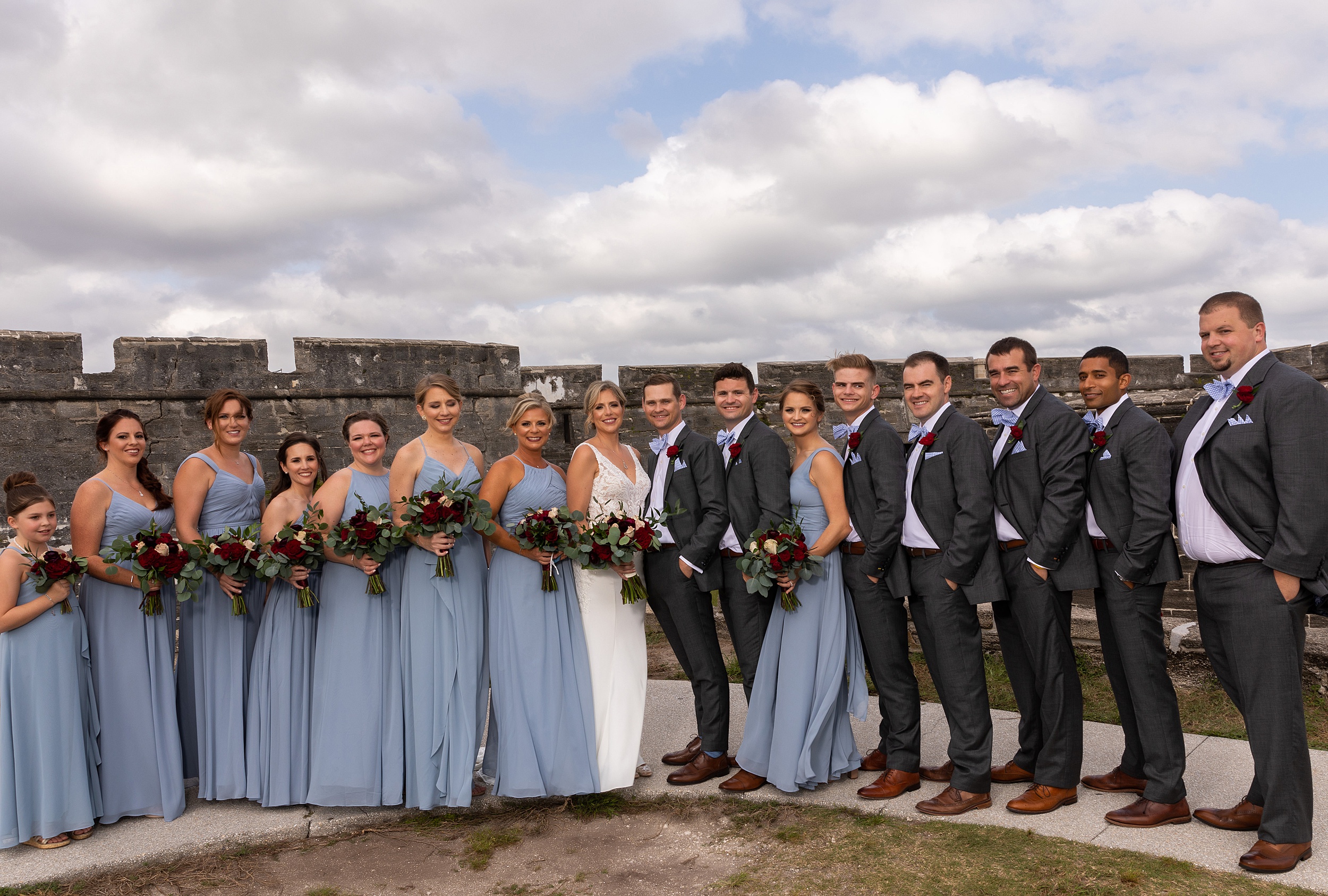 Newlyweds stand with their large wedding party by old walls on a sidewalk at their river house events wedding