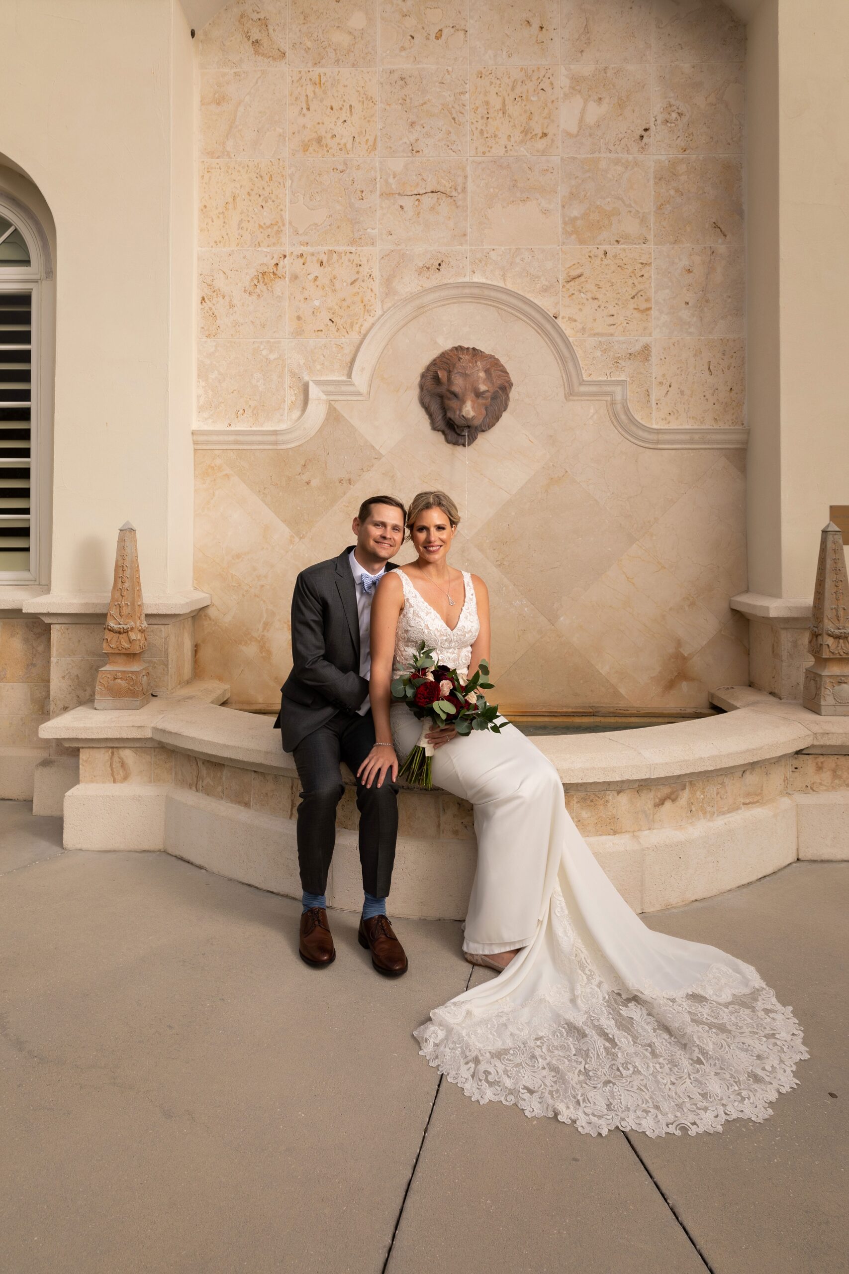 Newlyweds sit smiling on the edge of a large indoor marble fountain