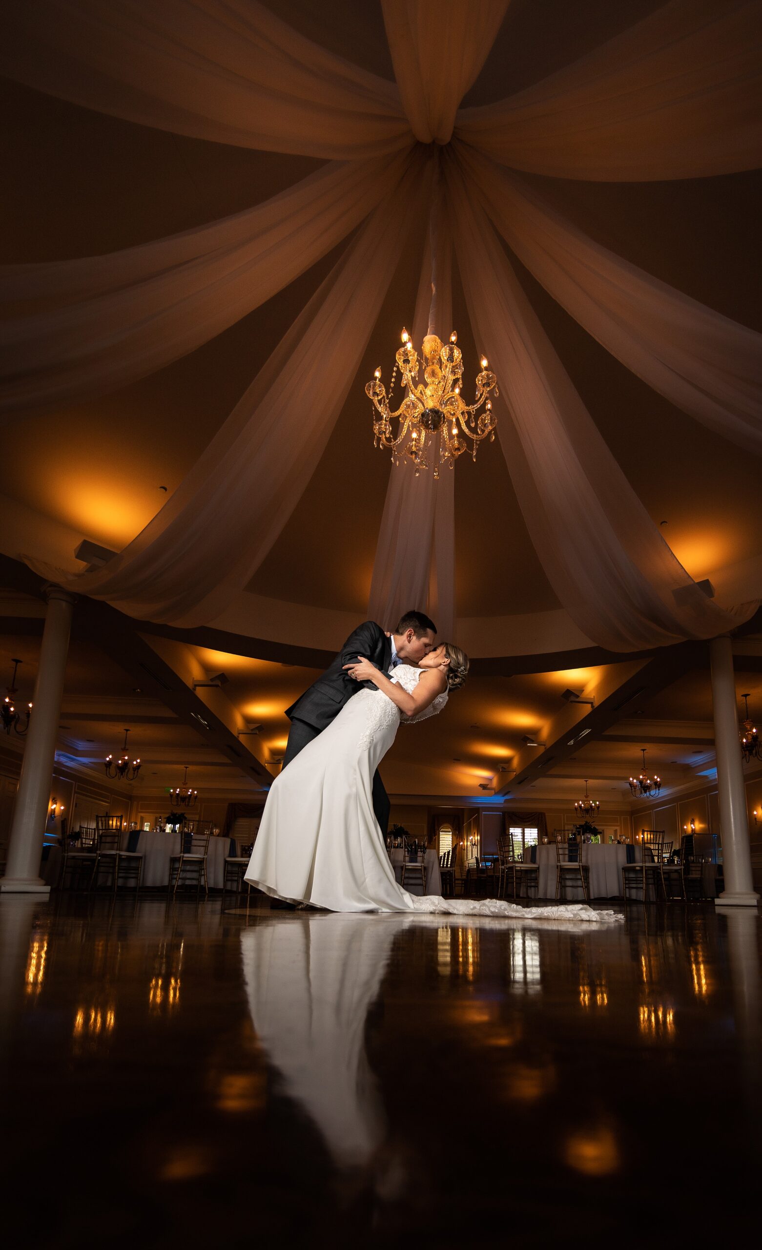 A groom dips and kisses his bride underneath a crystal chandelier in the center of the river house events wedding reception dance floor