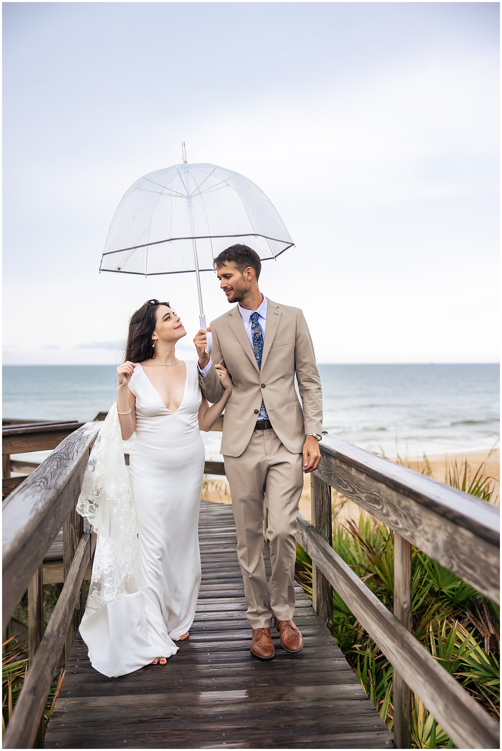 Newlyweds walk under a clear umbrella over a wet beach boardwalk