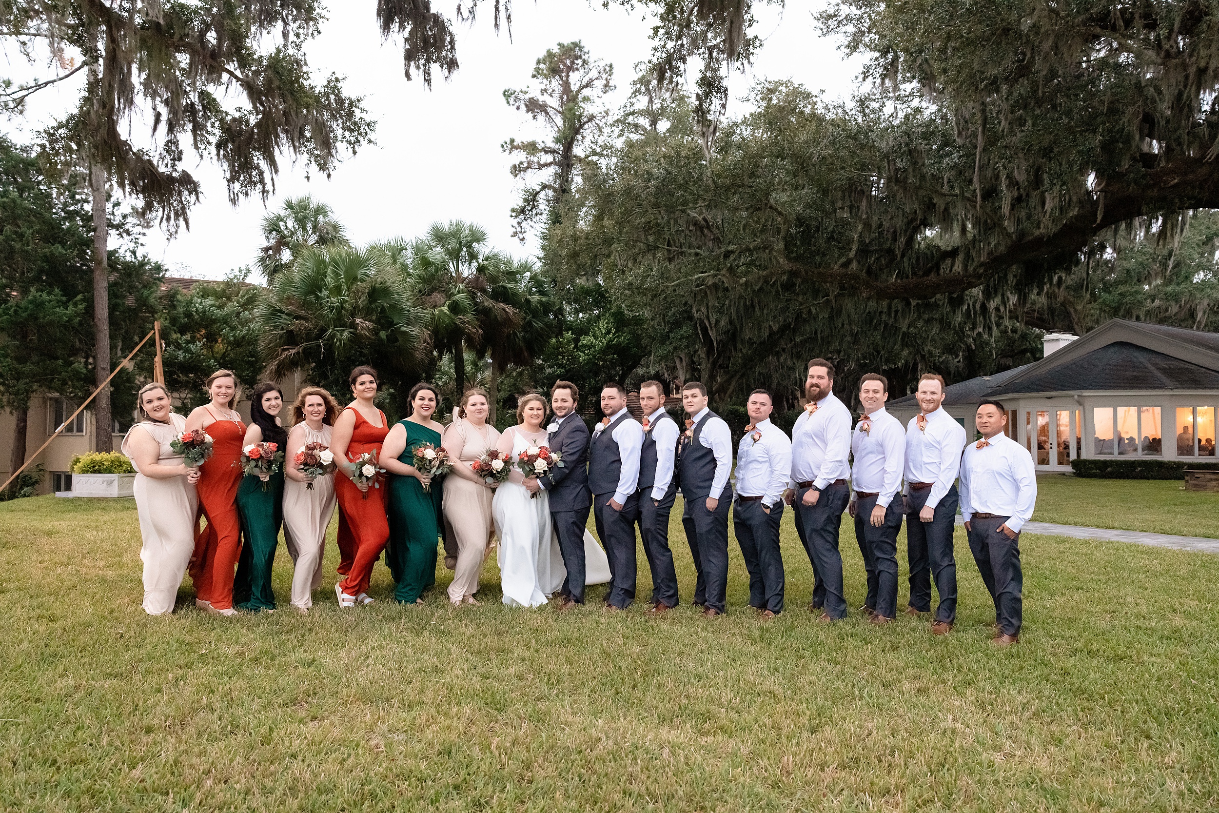 A large wedding party stands with the bride and groom on a windy lawn under oak trees at a bok tower gardens wedding