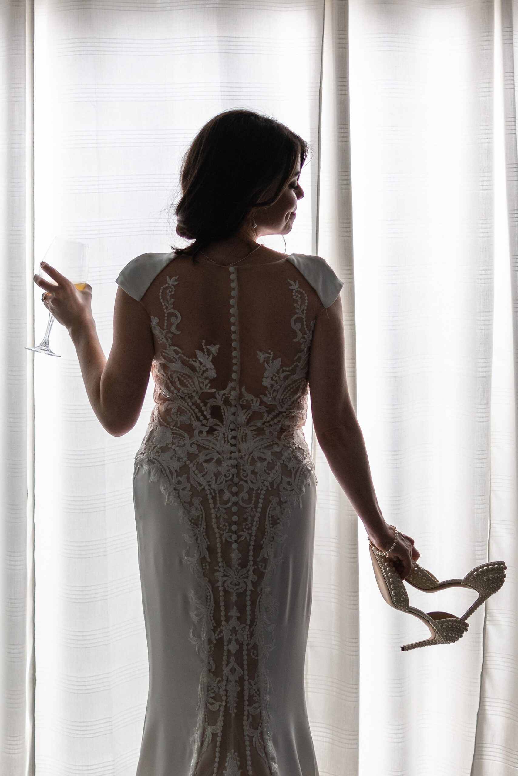 A bride stands in a window showing off her elaborate lace embroidery and holding a wine glass and shoes