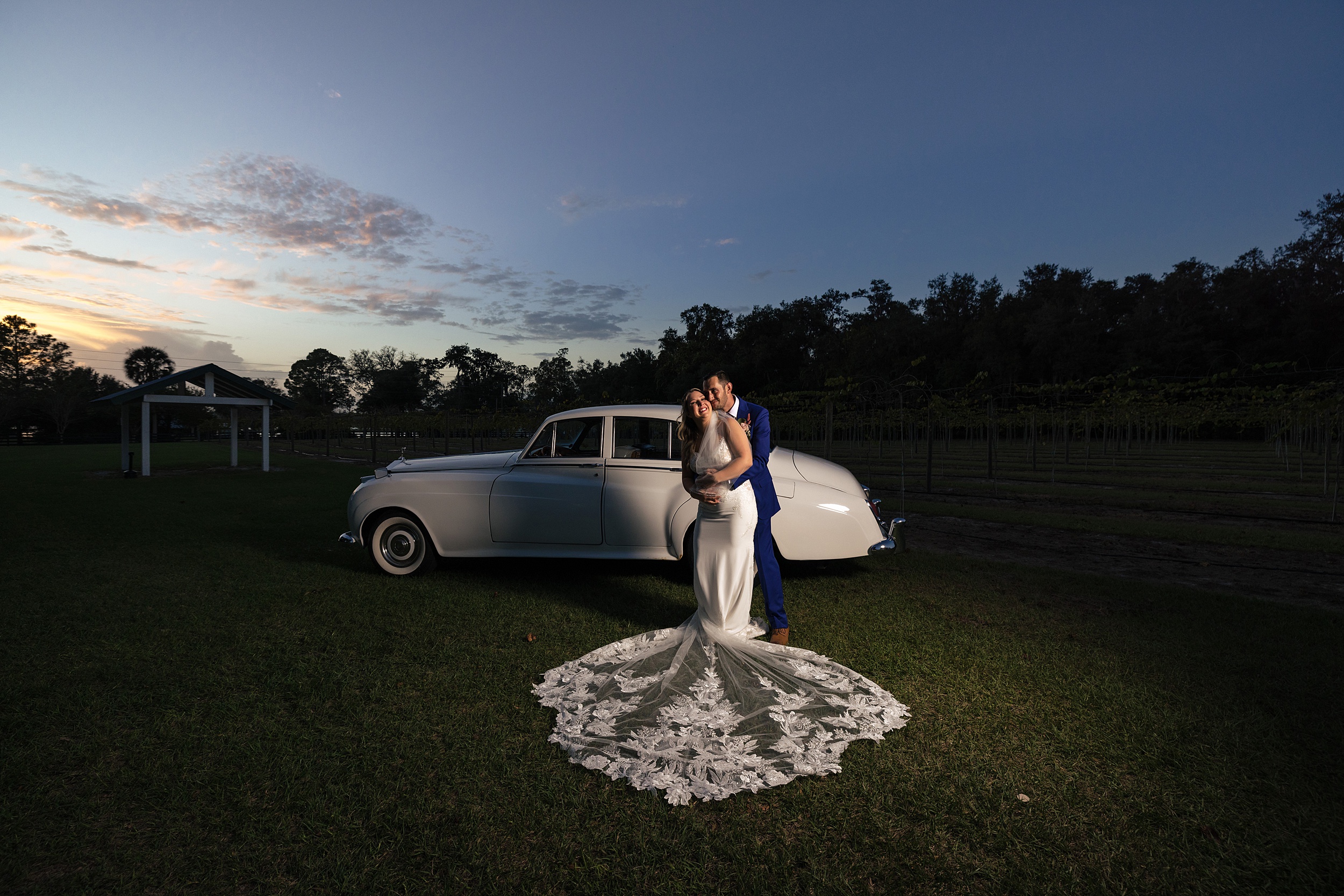 Newlyweds laugh together in a hug while standing in a field with a vintage car at sunset Ever After Farms Weddings