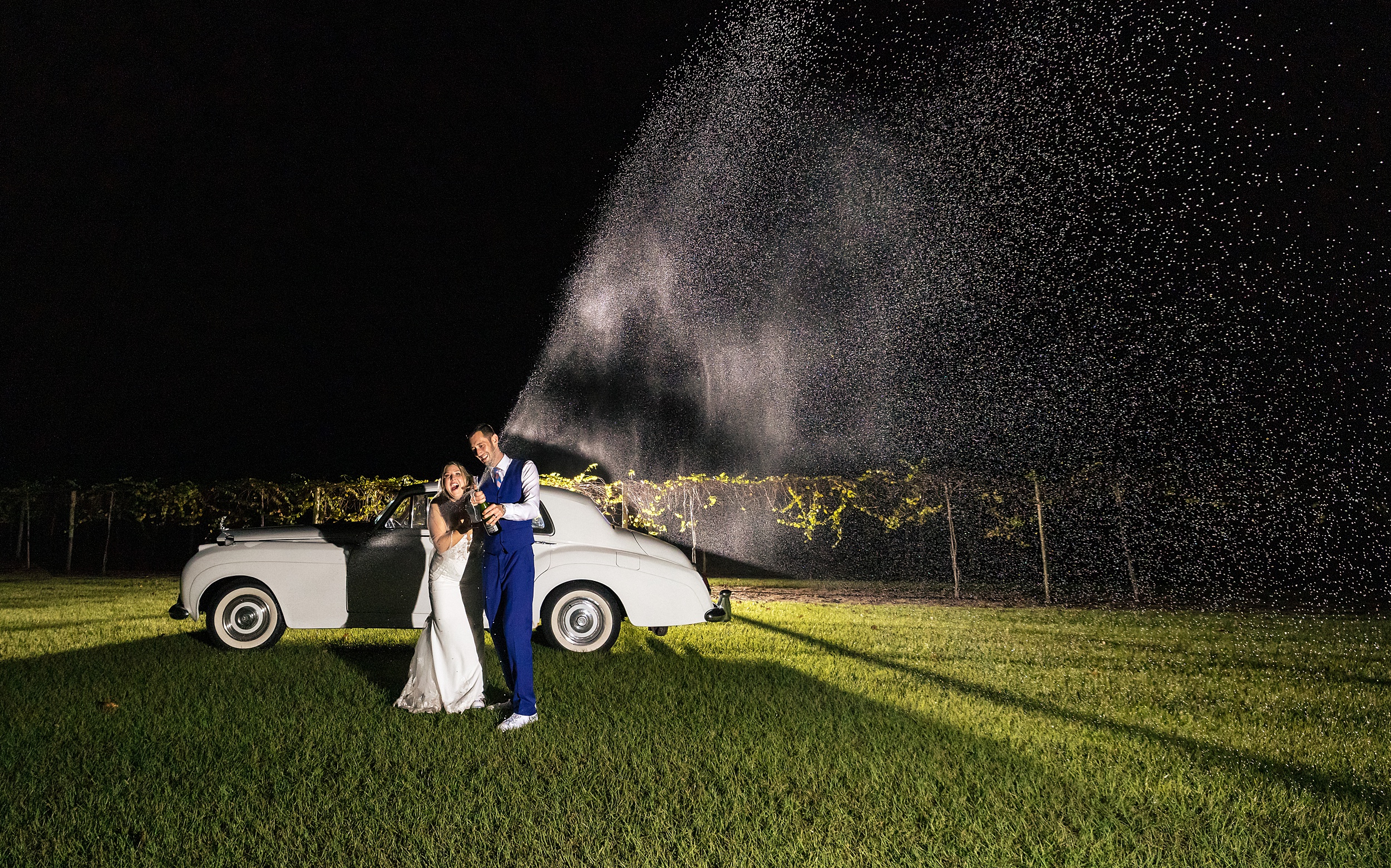 Newlyweds pop champagne while standing in a lawn with a vintage white car