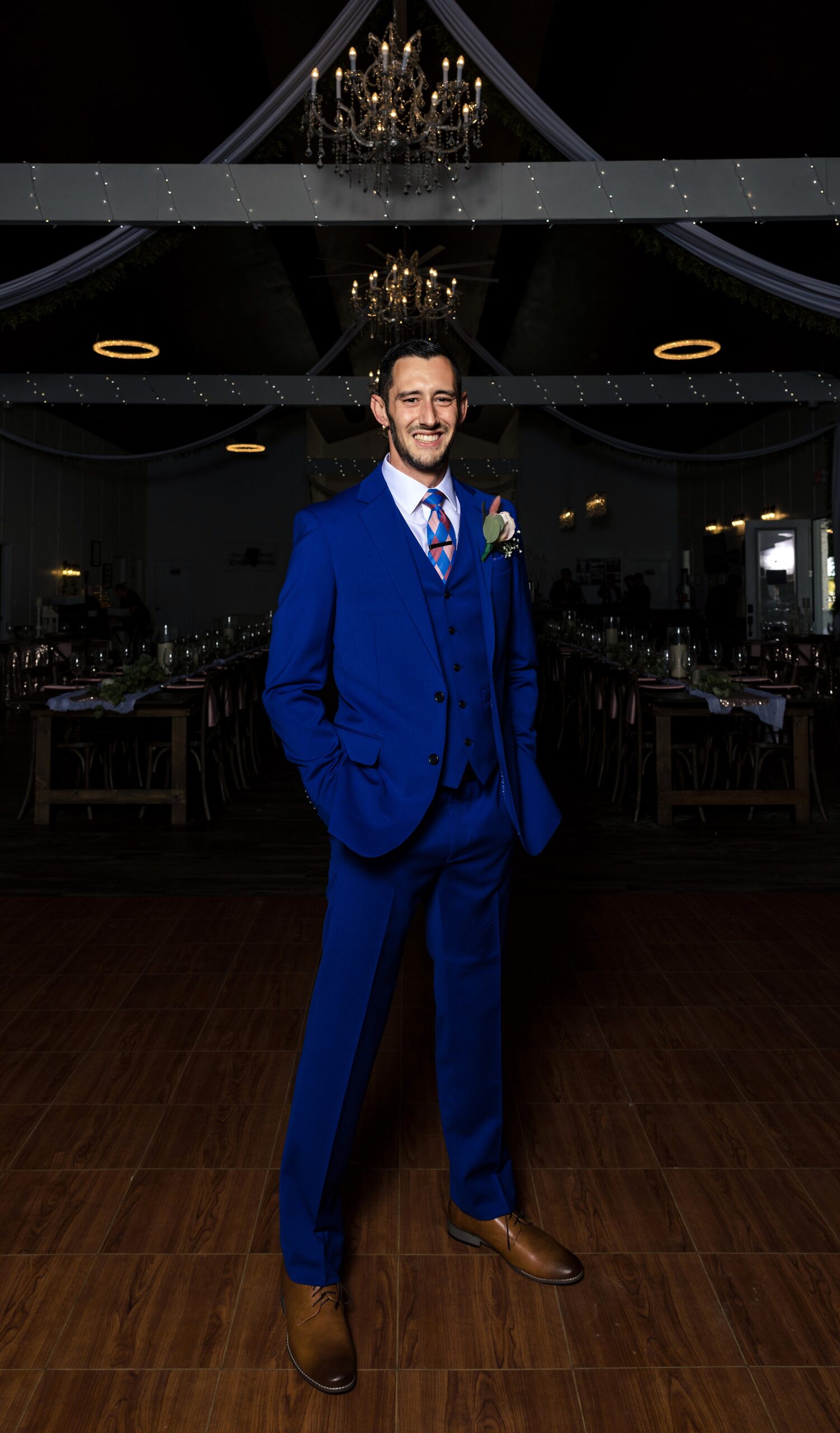 A groom in a blue suit standing under chandeliers with hands in pockets