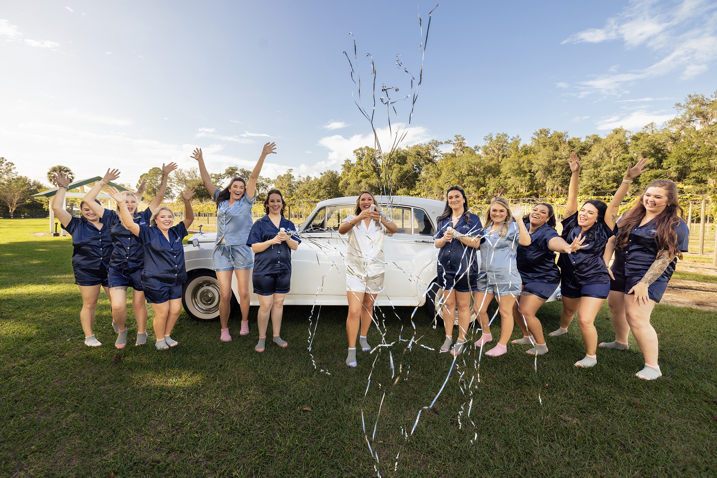 A bride and her bridal party pop streamers in a lawn with a white antique car Ever After Farms Weddings