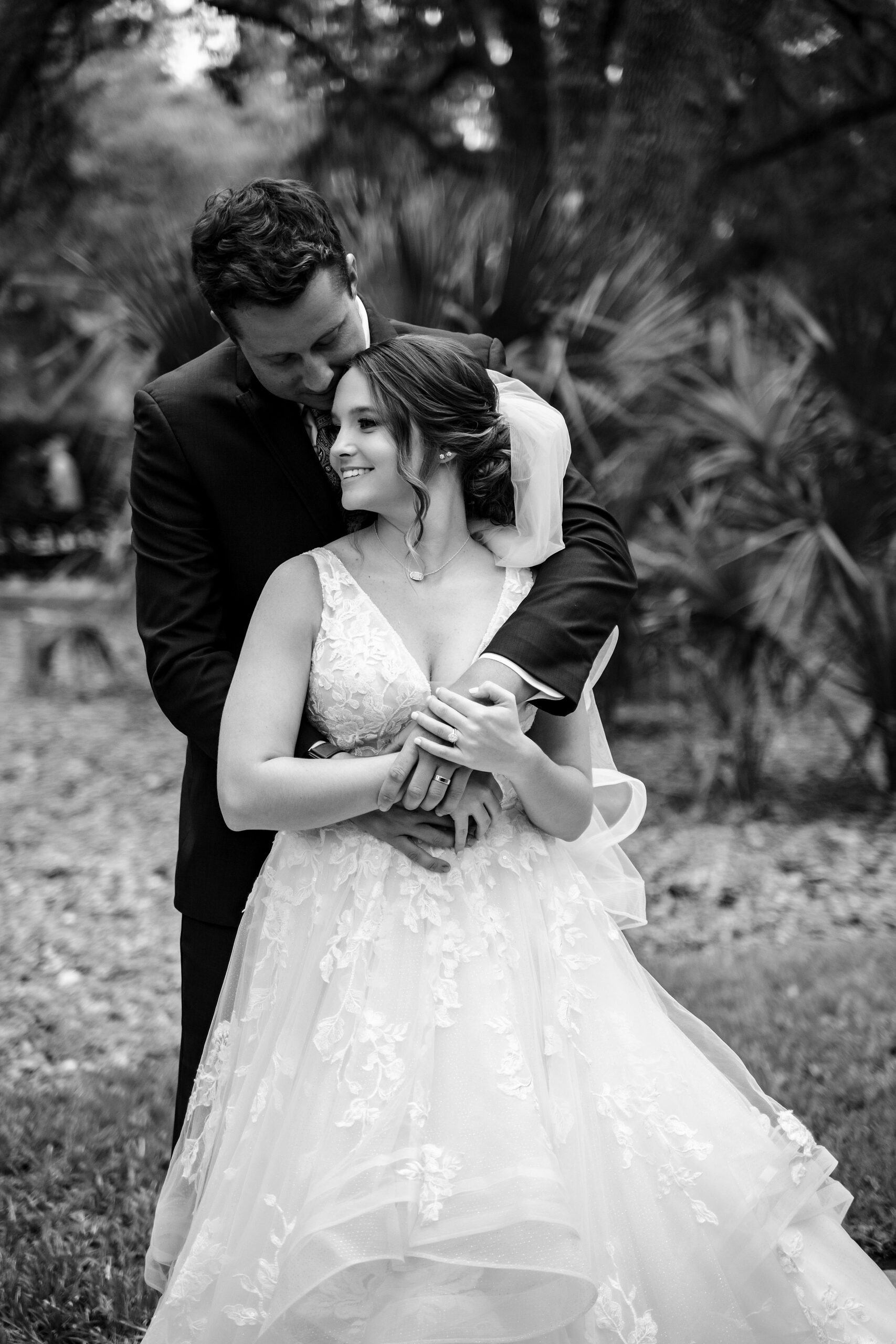 A groom hugs his bride from behind in a tropical garden while smiling