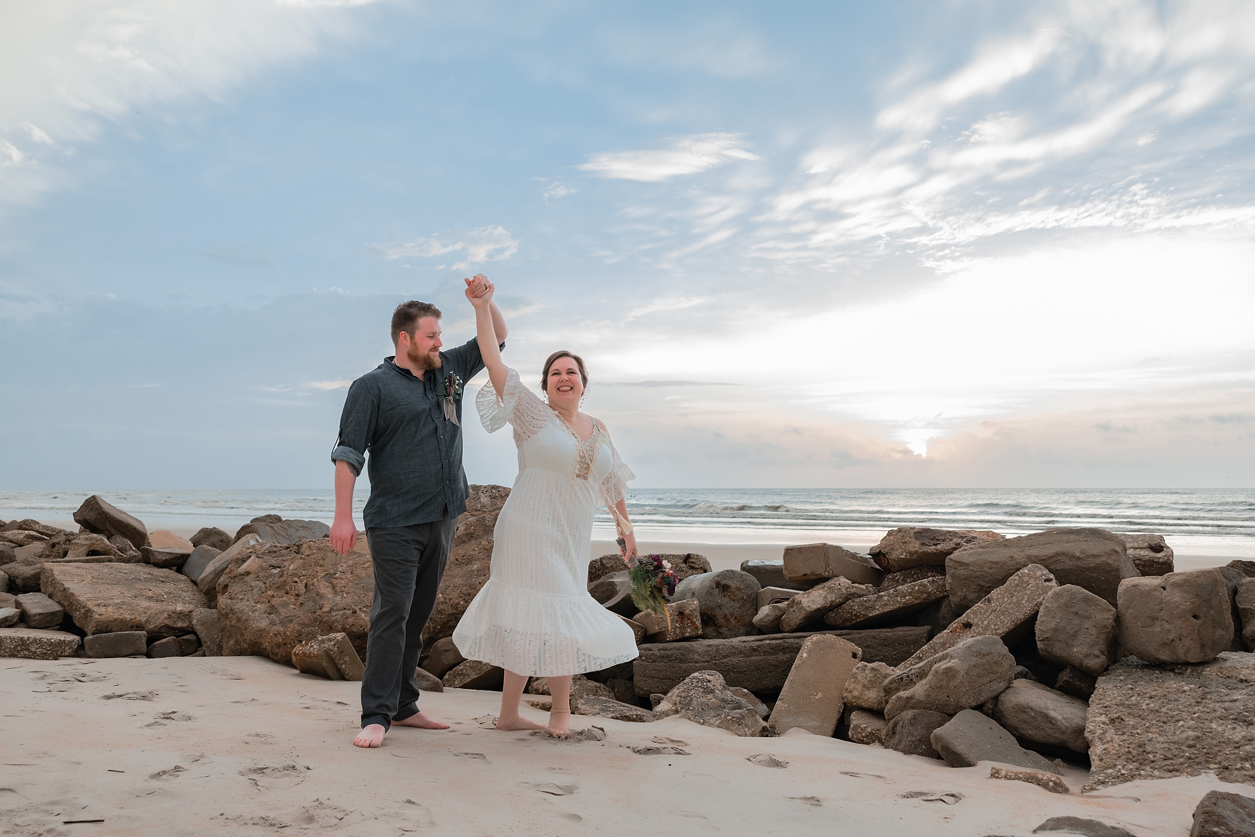 A groom twirls his bride while standing on a beach by some large rocks