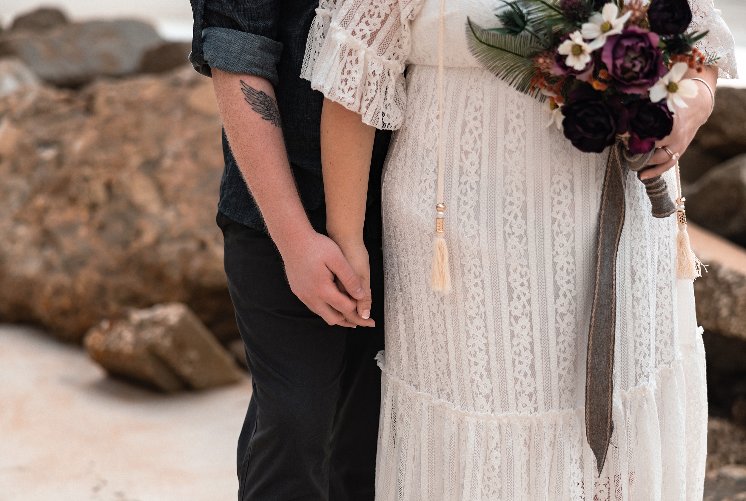 Details of newlyweds holding hands on a beach with a dark rose bouquet