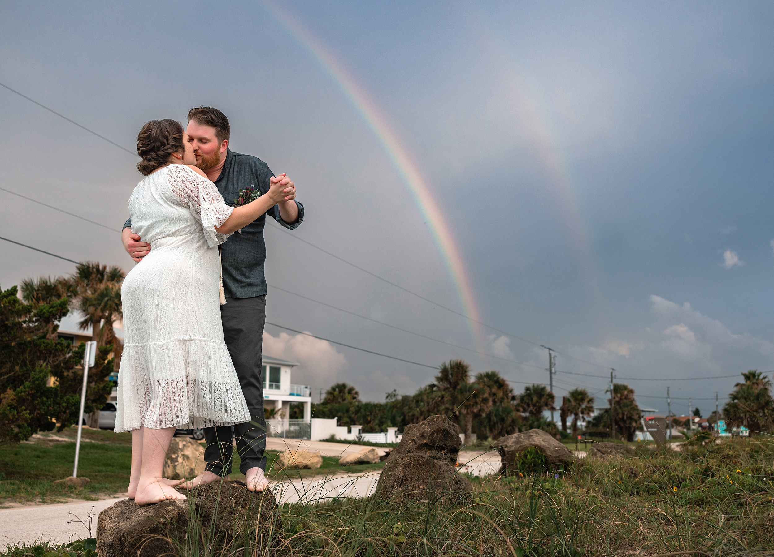 Newlyweds kiss while standing under a double rainbow on limestone rocks at the beach at their st augustine beach wedding venues