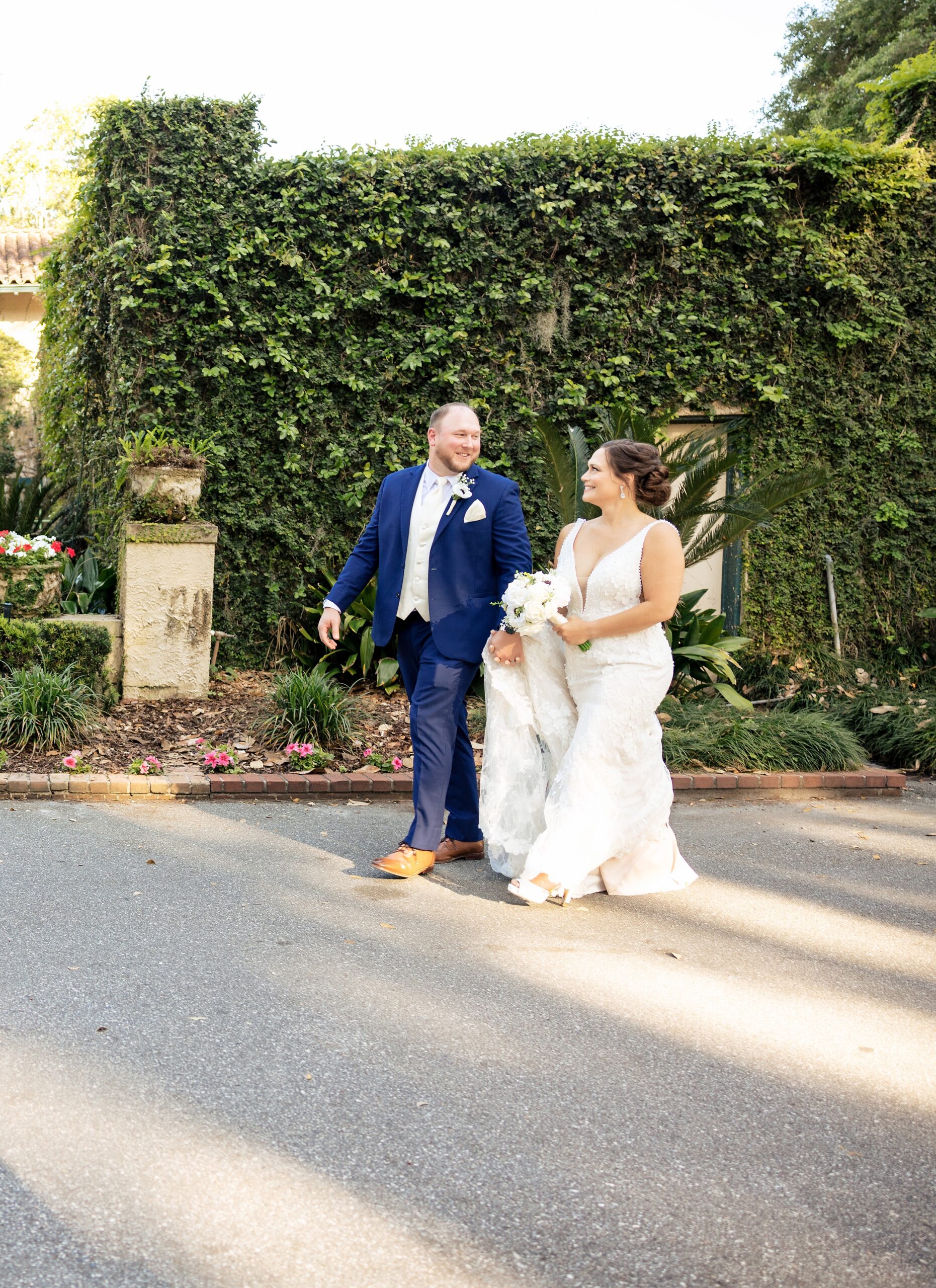 A groom holds the hand of his bride as they walk through a garden