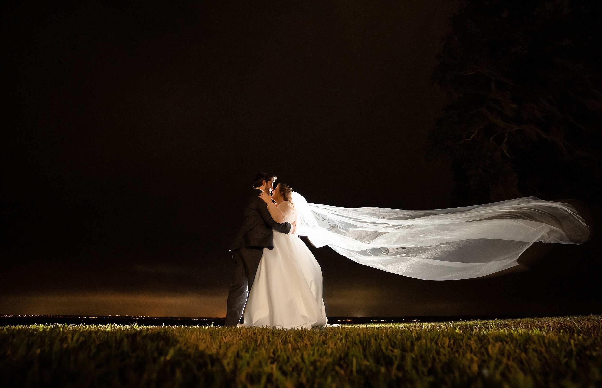 Newlyweds stand on a lawn at night kissing with the veil flowing long behind them in the wind