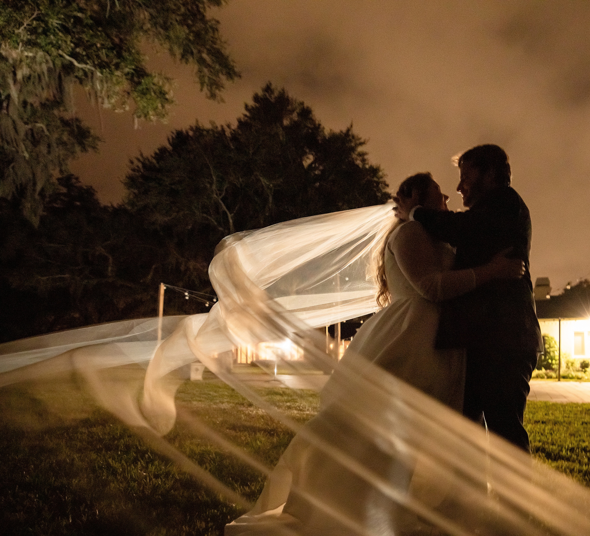 Newlyweds embrace for a kiss while standing in a lawn at night with the veil flowing around them at their the enchanting barn wedding