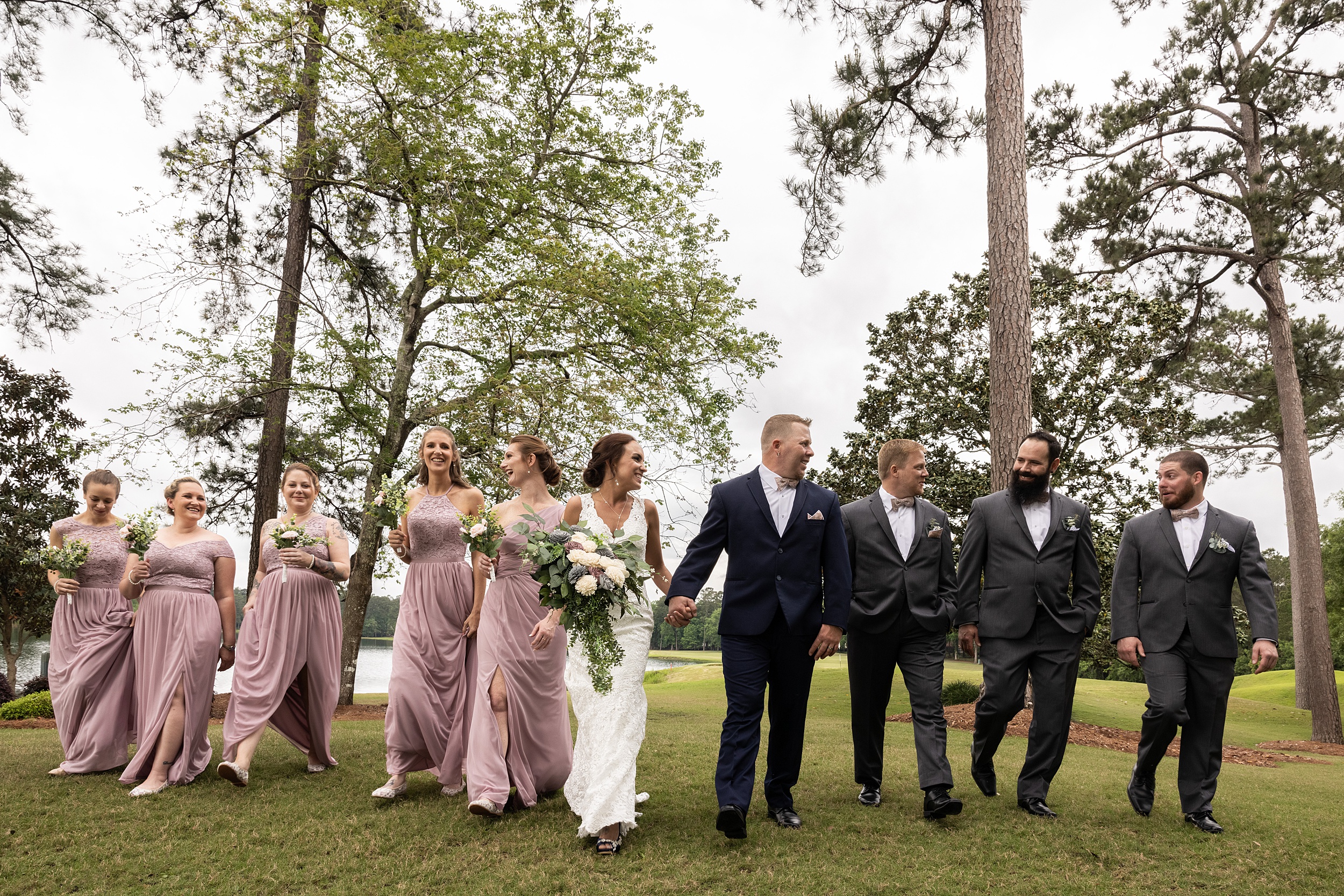 Newlyweds walk hand in hand through a green field under trees with their bridal party at their the howey mansion wedding