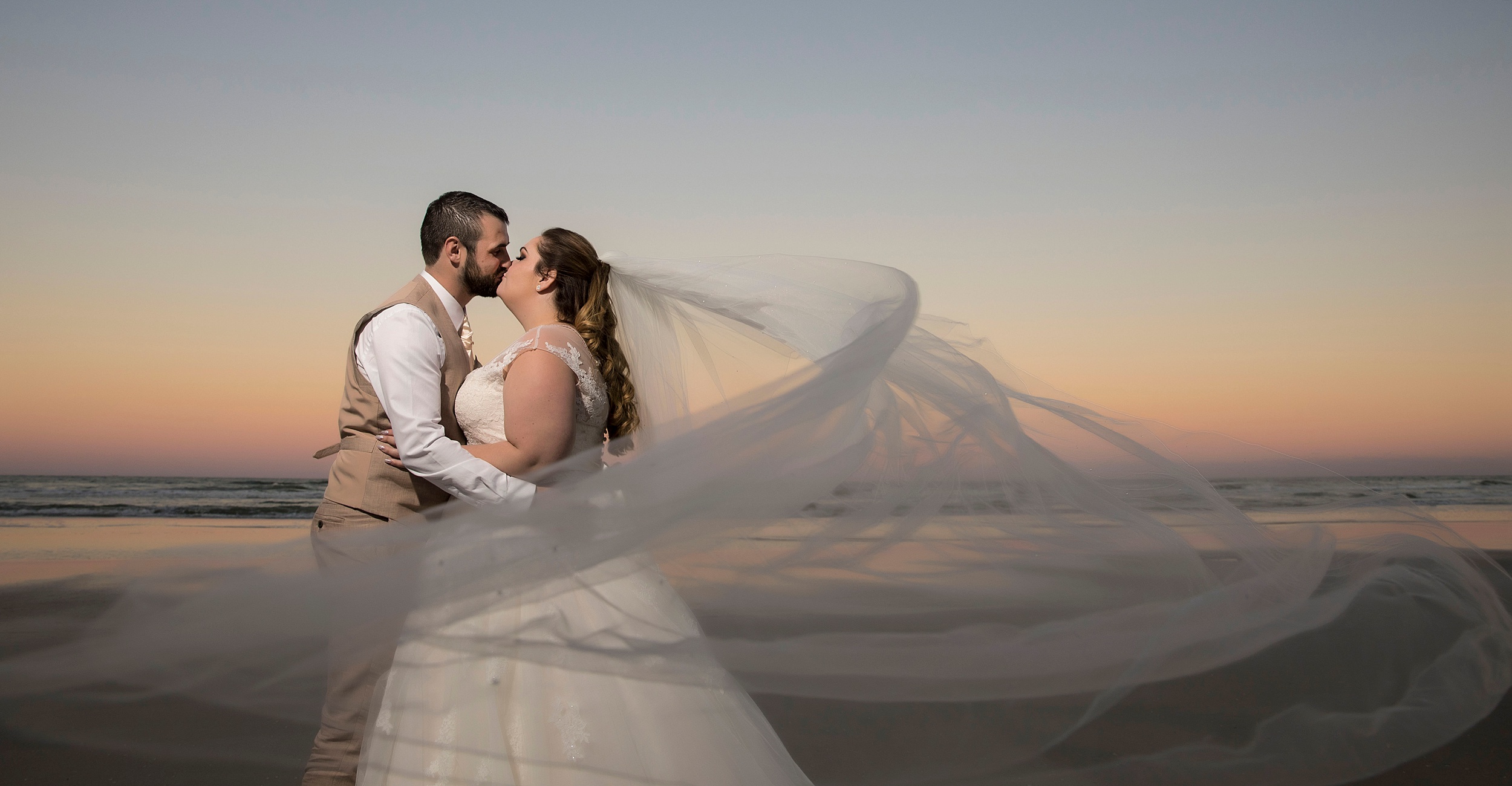 Newlyweds kiss at sunset on the beach as the veil flows in the wind around them