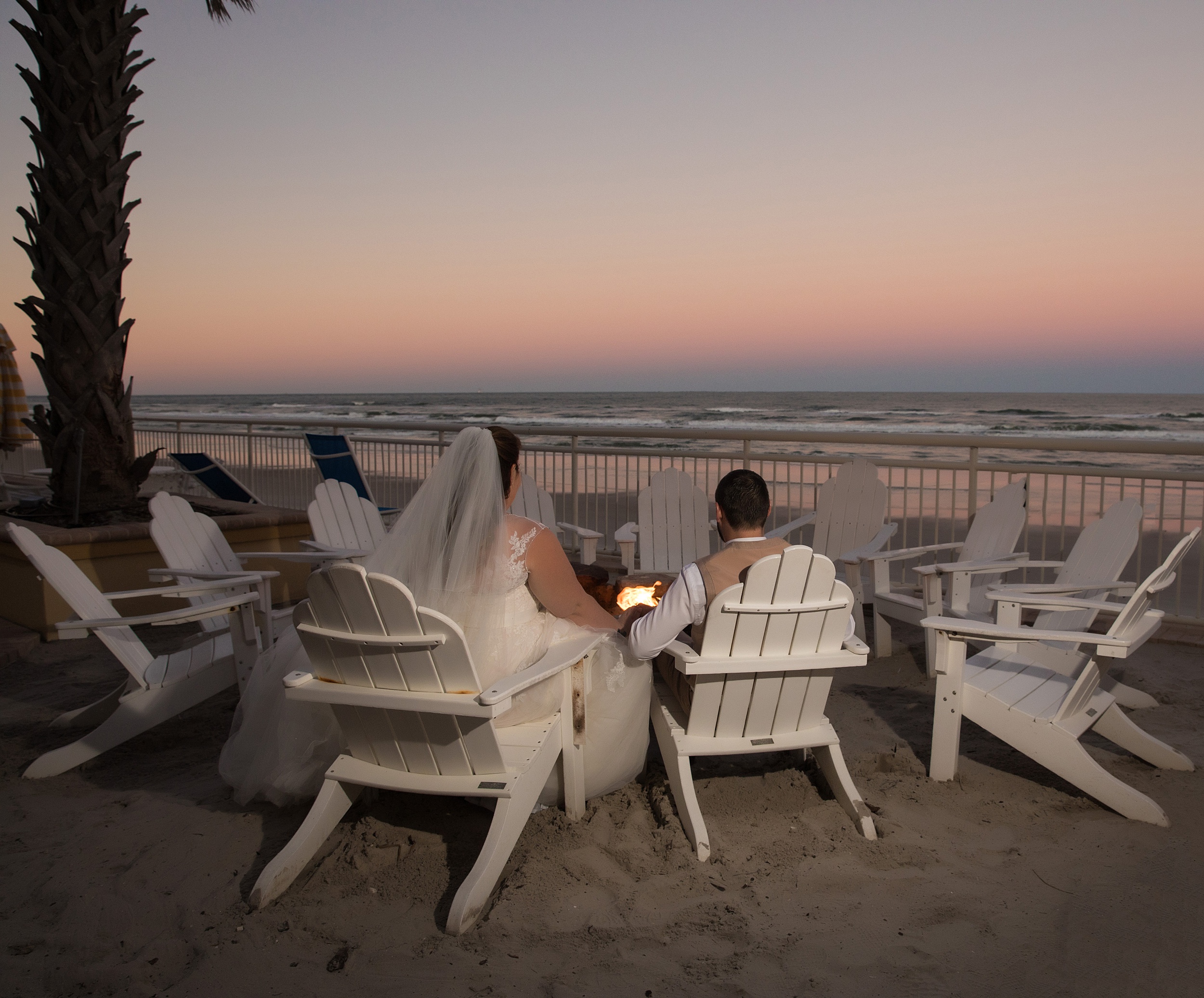 Newlyweds sit by the fire in Adirondack chairs on the beach
