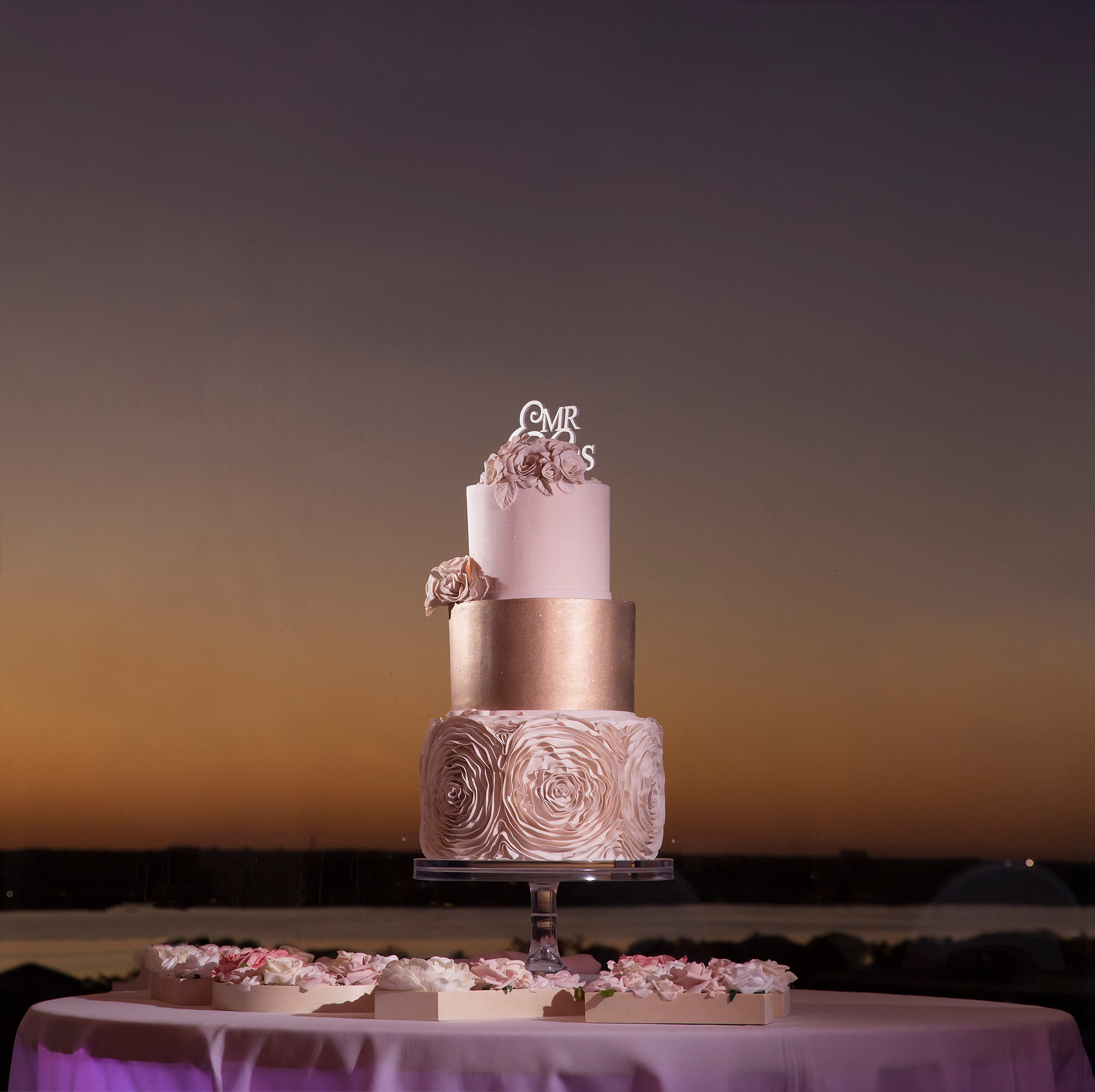 A three tier wedding cake sits on a beachfront patio table at sunset