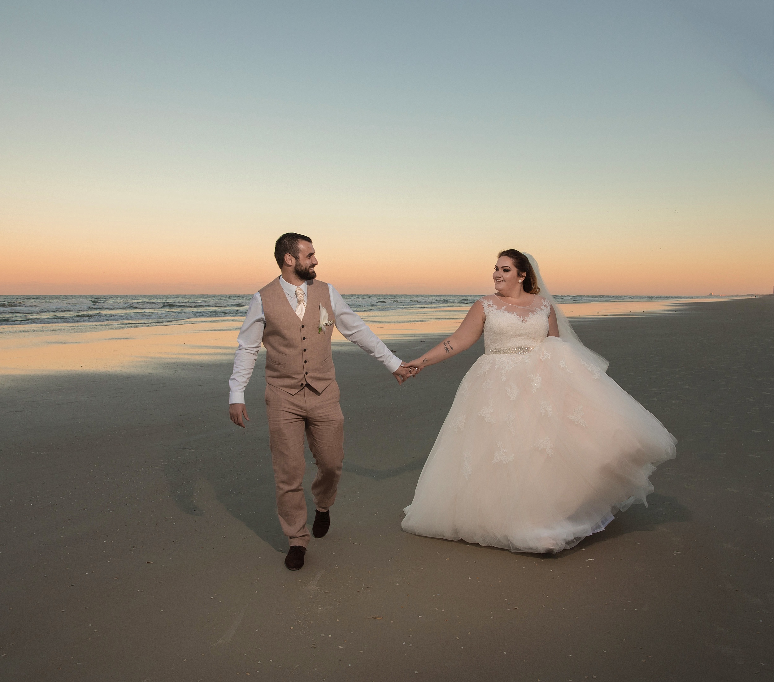A groom in a tan suit leads his bride by the hand up a beach at sunset at their the shores resort and spa wedding
