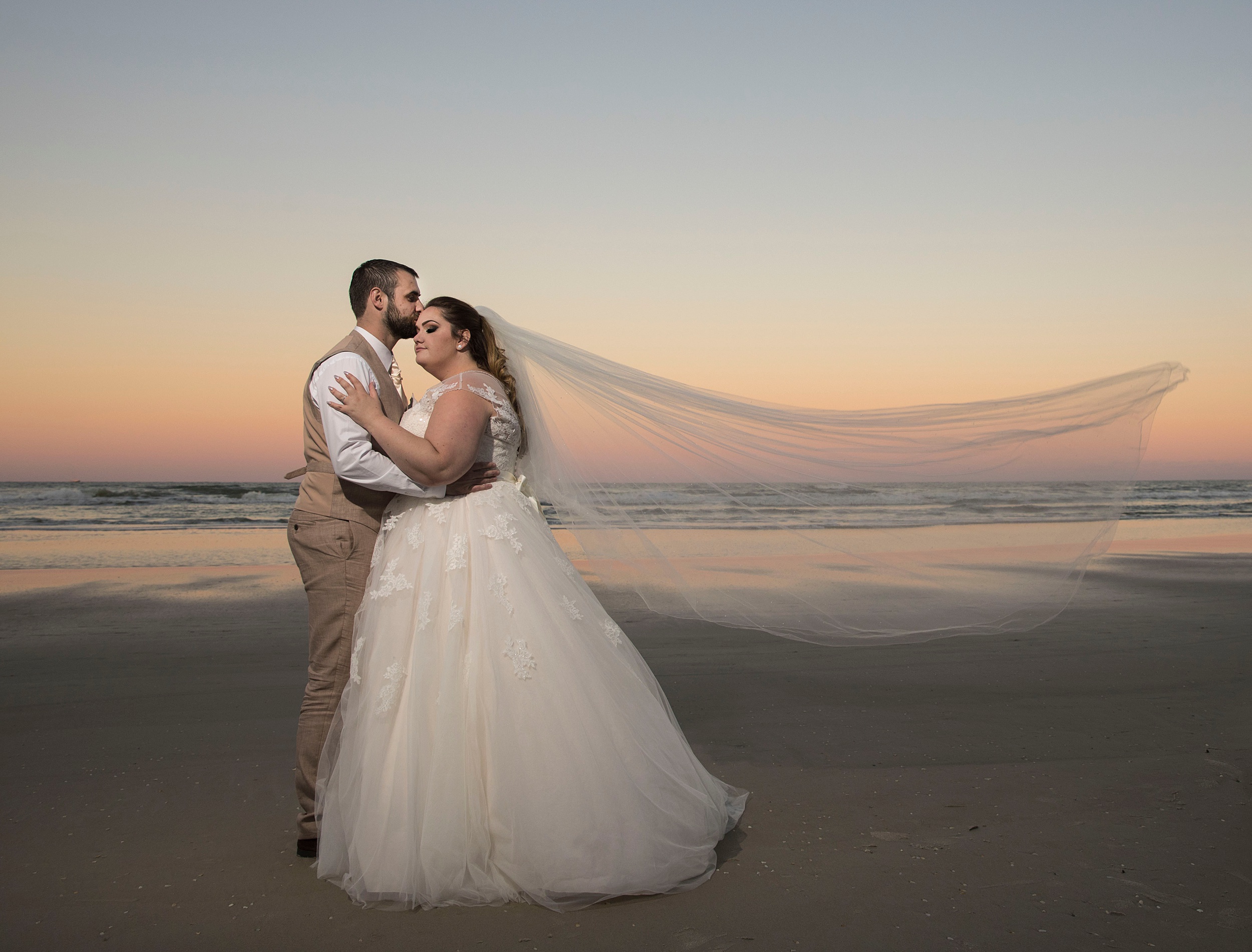 Newlyweds kiss while standing on the beach at sunset as the veil blows in the wind during their the shores resort and spa wedding