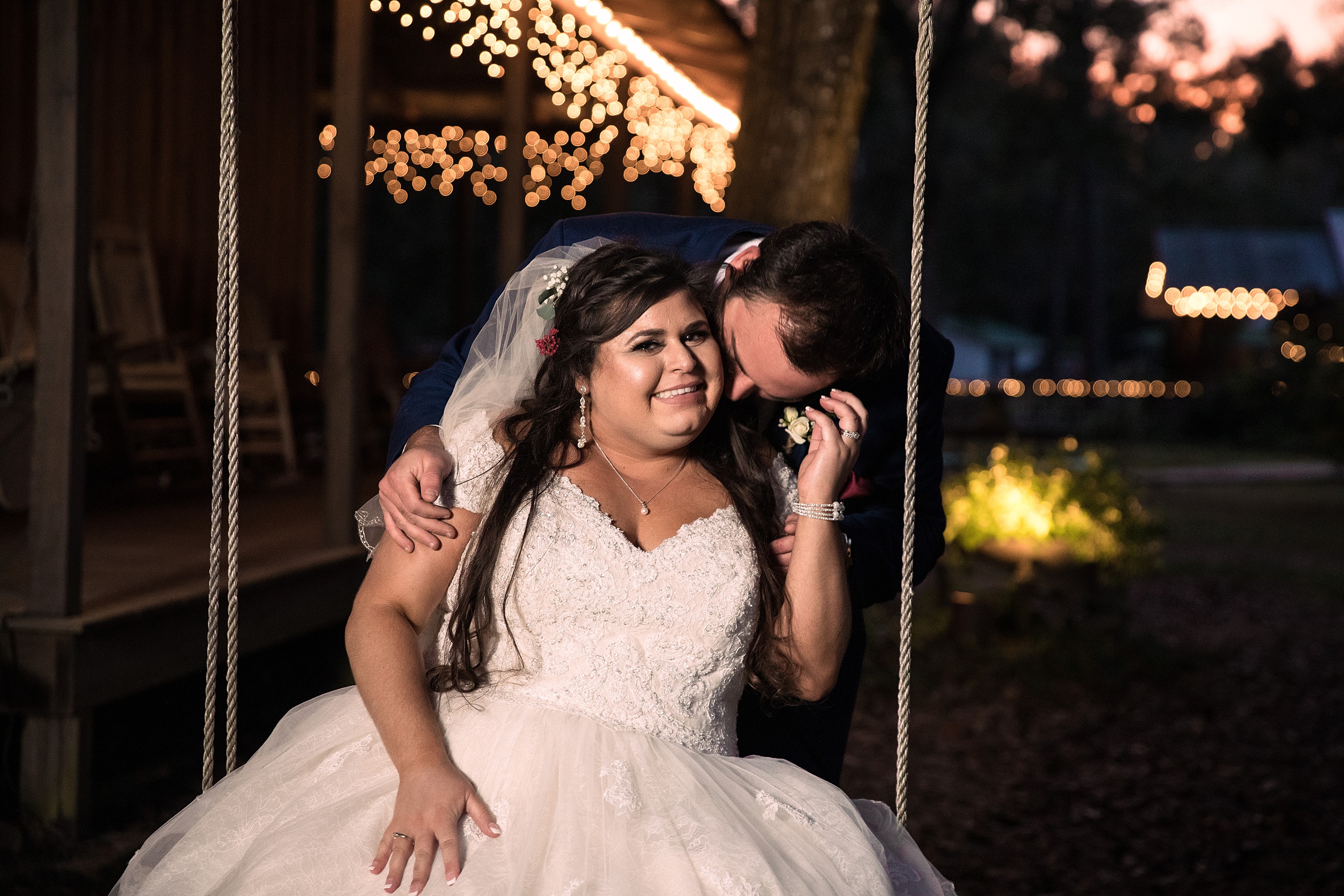 A groom kisses his bride's cheek as she sits on a swing by a porch under market lights