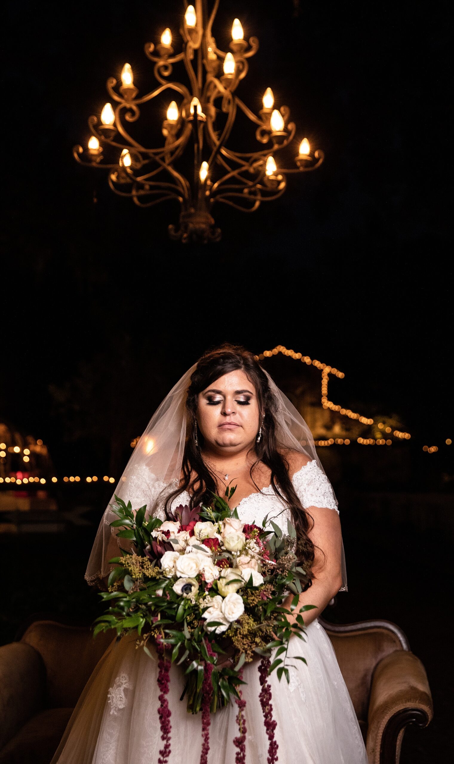A bride stands with eyes closed under an outdoor chandelier holding her colorful large bouquet