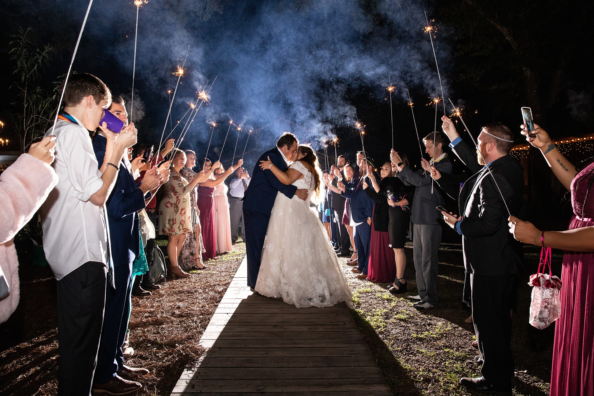 Newlyweds kiss while exiting their reception at night surrounded by guests holding sparklers